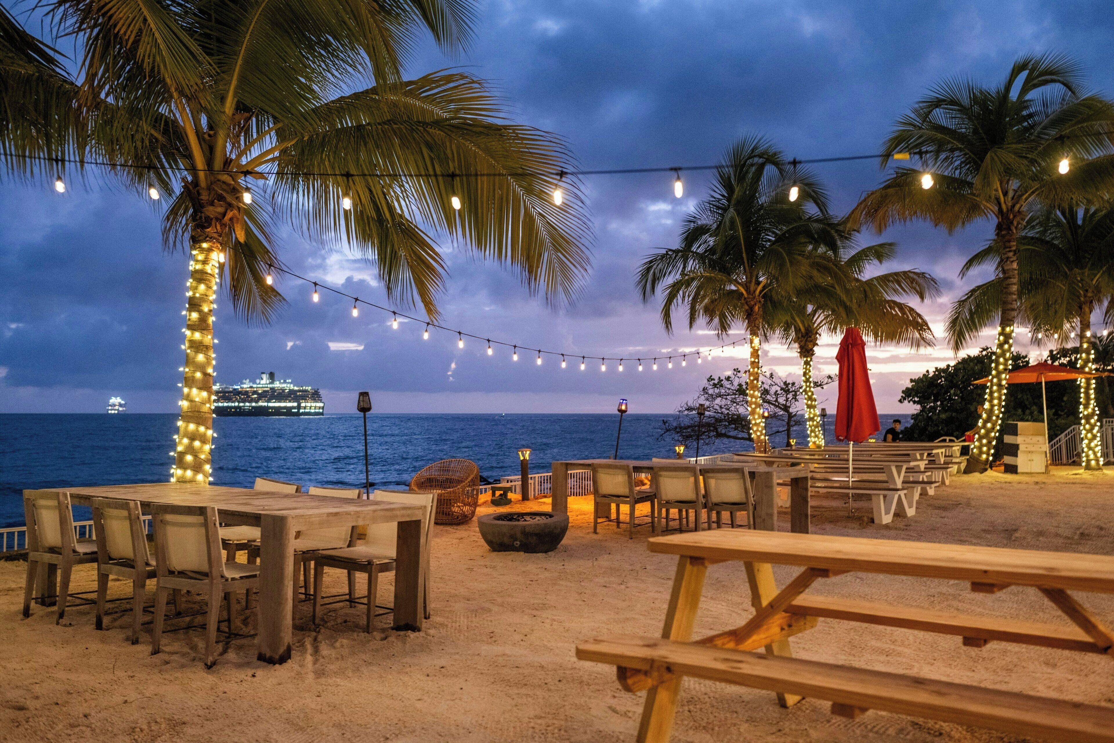 A beachside dining area with string lights, palm trees, and a view of the ocean.