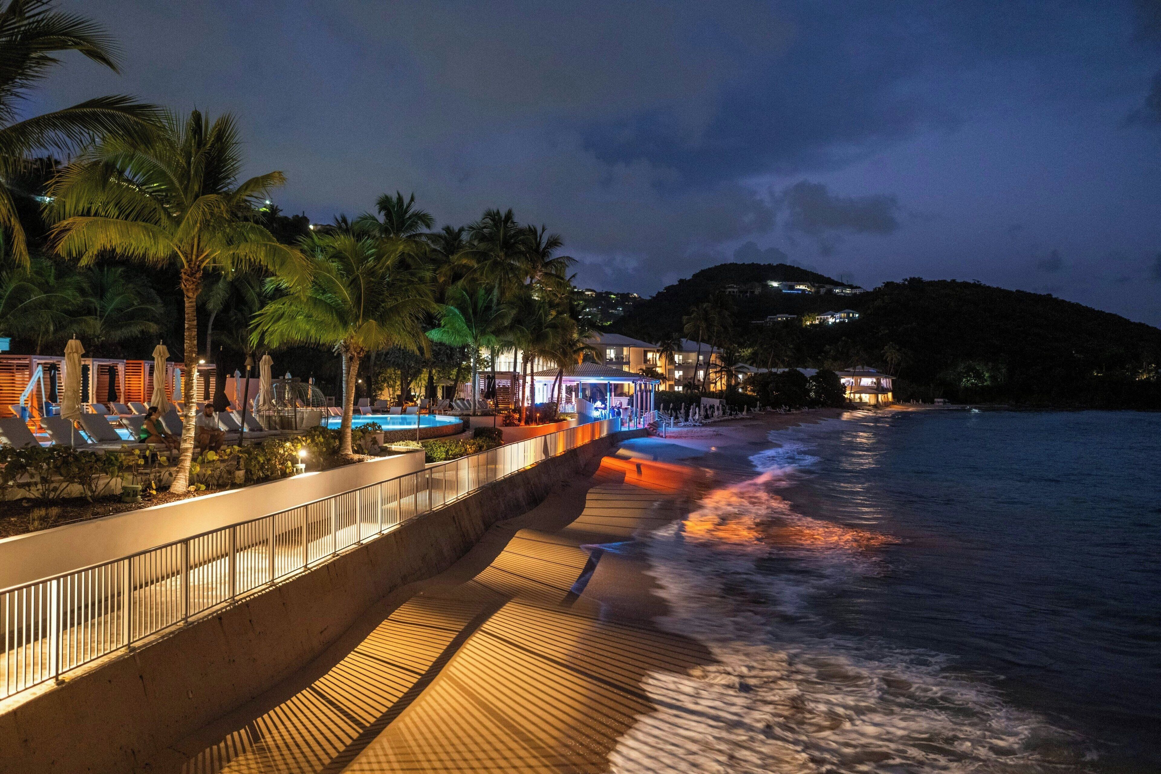 A beachfront hotel with a pool, palm trees, and a wooden walkway along the water.
