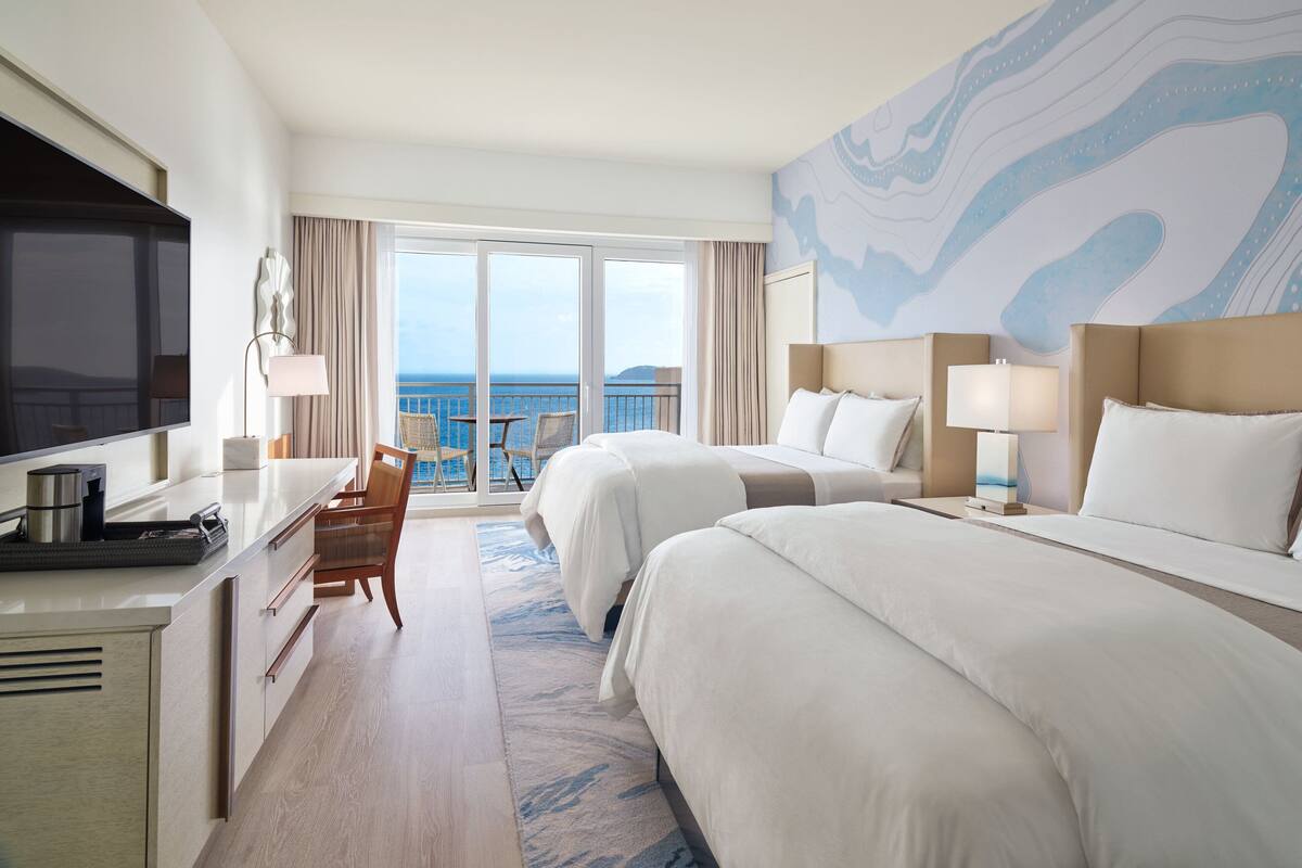 A hotel room with two beds, a TV, a desk, and a balcony with a view of the sea.