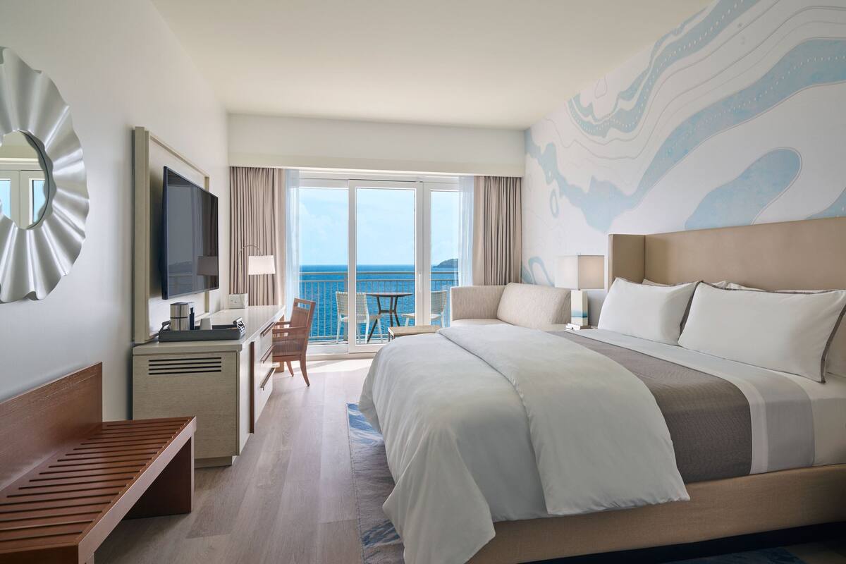 A hotel room with a large bed, a desk with a TV, a chair, and a view of the ocean.