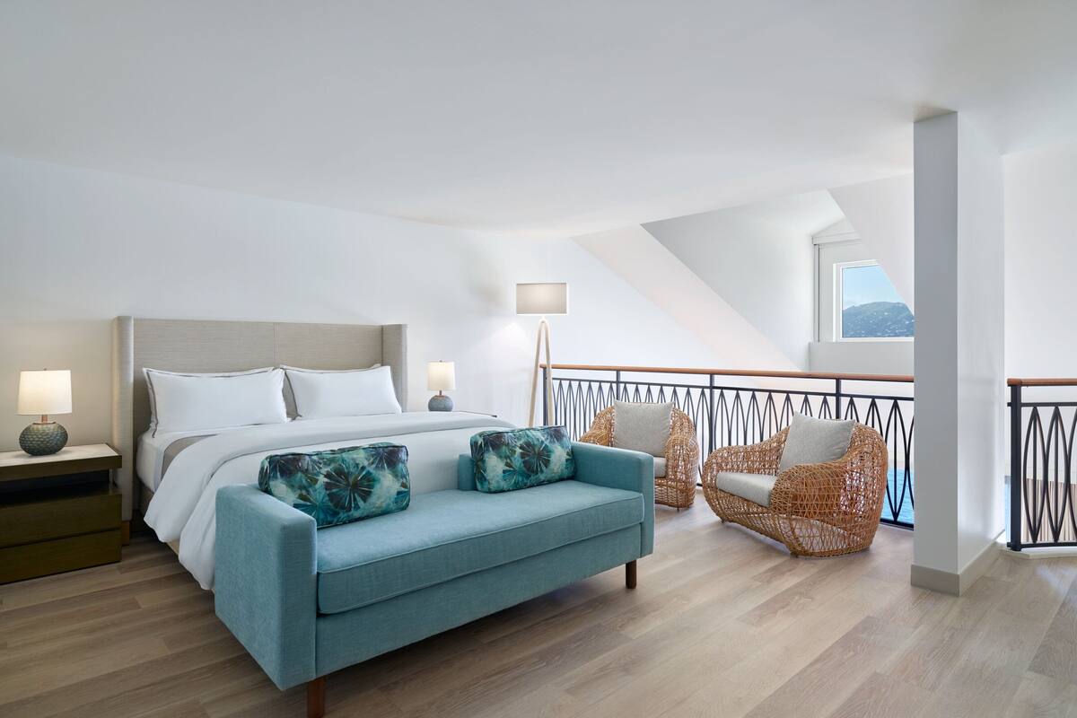 A modern bedroom with a bed, a blue sofa, wicker chairs, and a balcony with a railing.