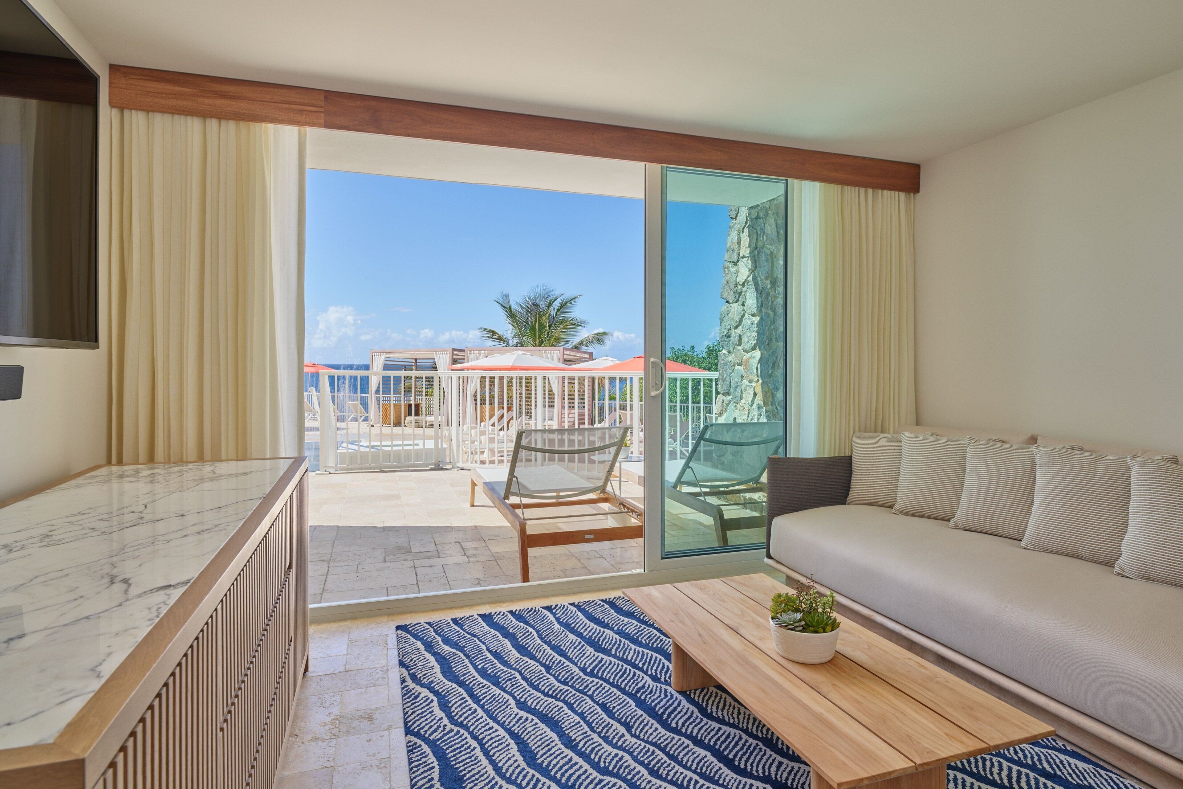 A modern hotel room with a marble countertop, a sofa, a coffee table with a plant, and a view of the outdoor area with a pool and lounge chairs.
