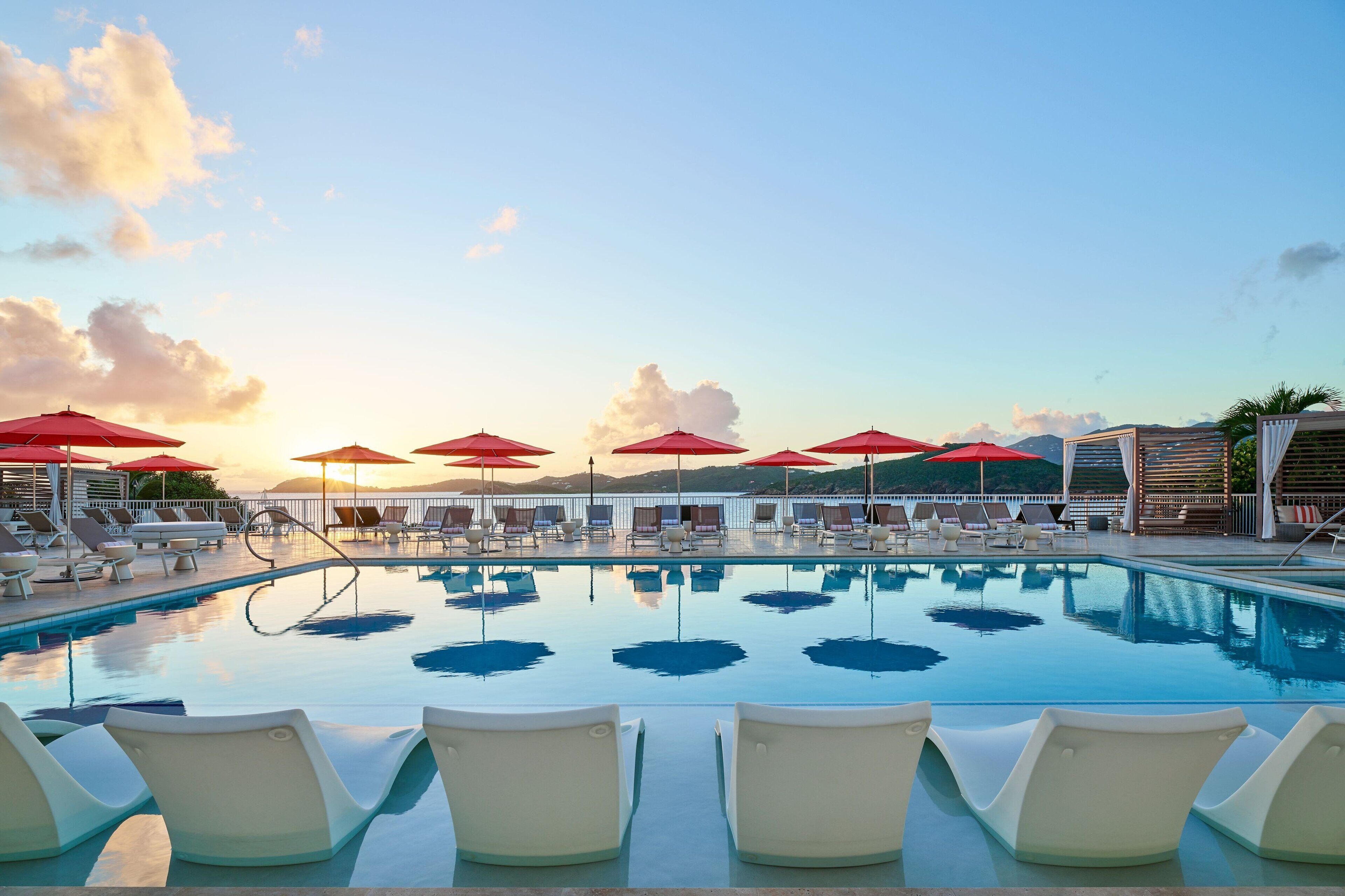 A pool area with lounge chairs and red umbrellas, surrounded by a pool with clear water and a view of mountains in the background.
