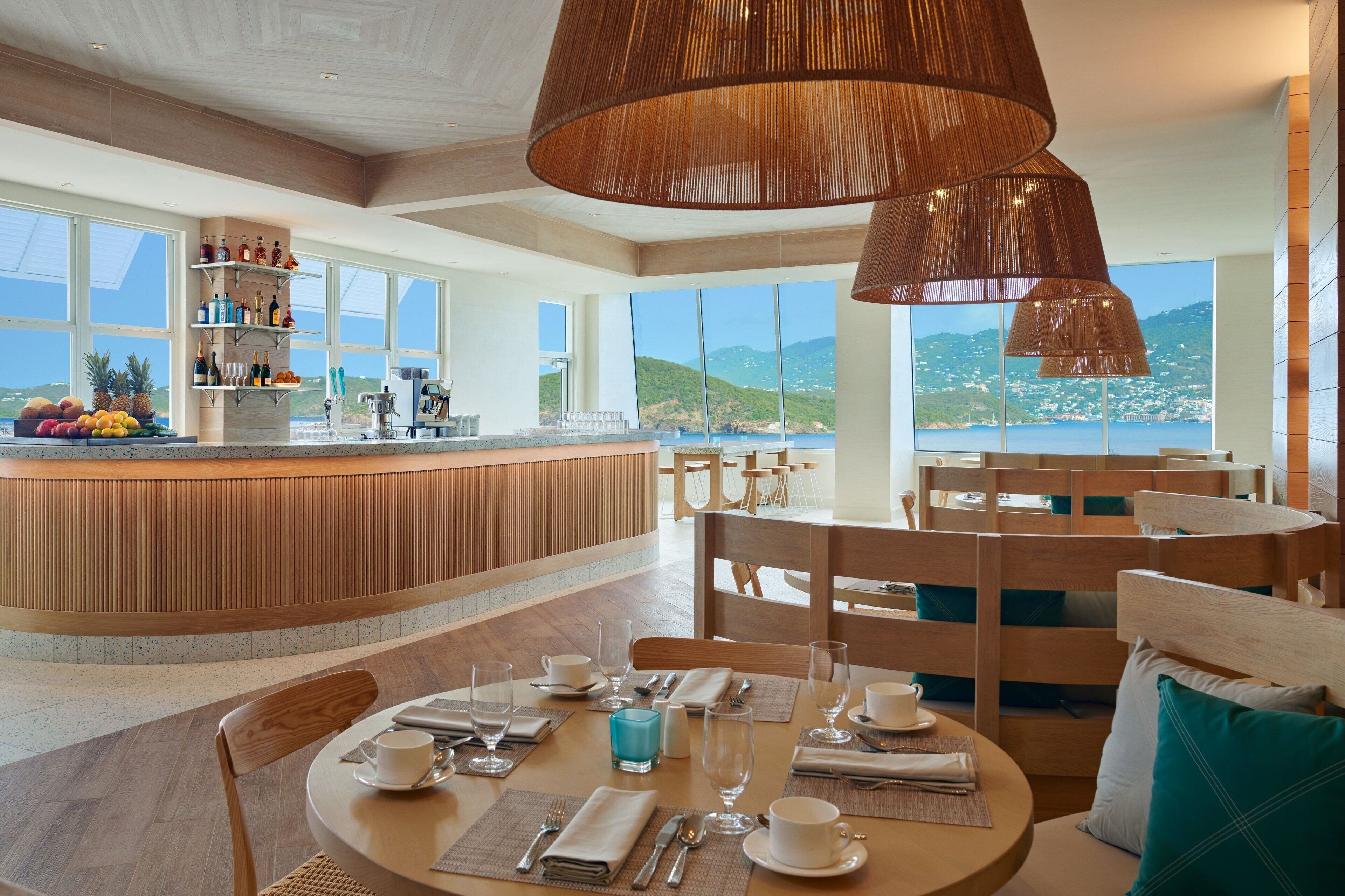 A restaurant with a wooden counter and seating area, large windows with a view of the sea and hills, and hanging pendant lights.