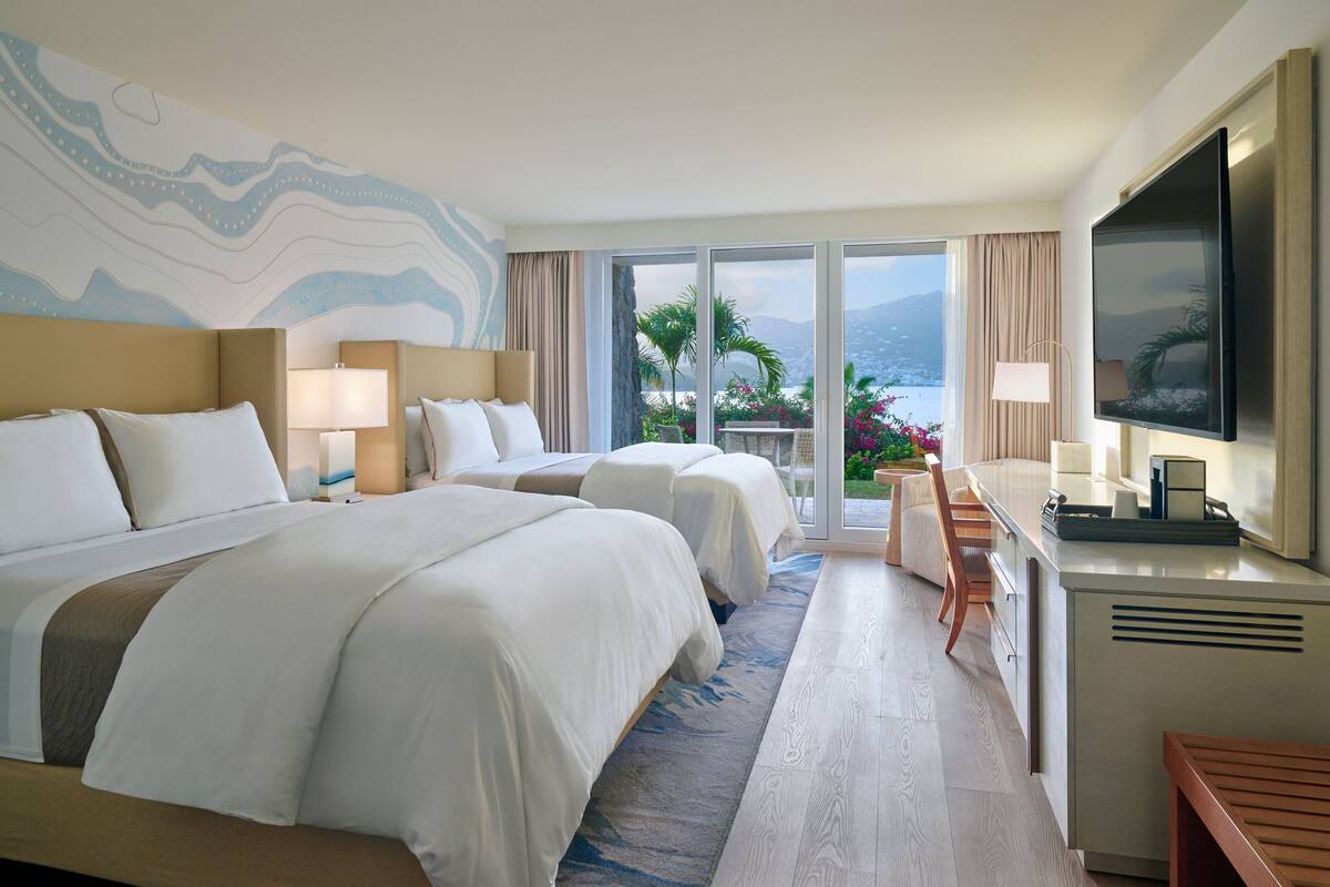 A hotel room with two beds, a TV, a desk, and a balcony with a view.