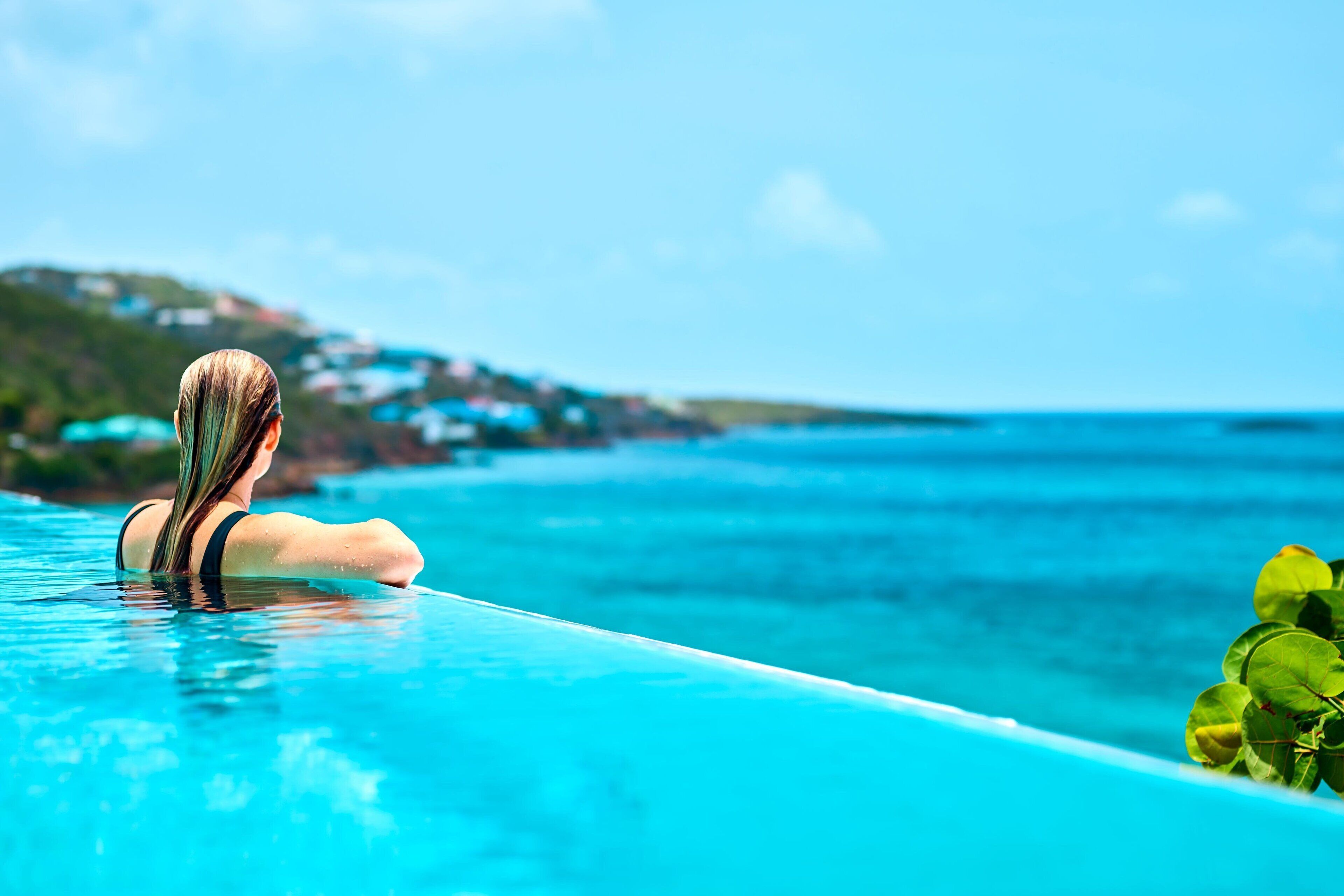 Una persona relajándose en una piscina infinita de color azul cristalino, con vistas al océano y a una costa lejana.