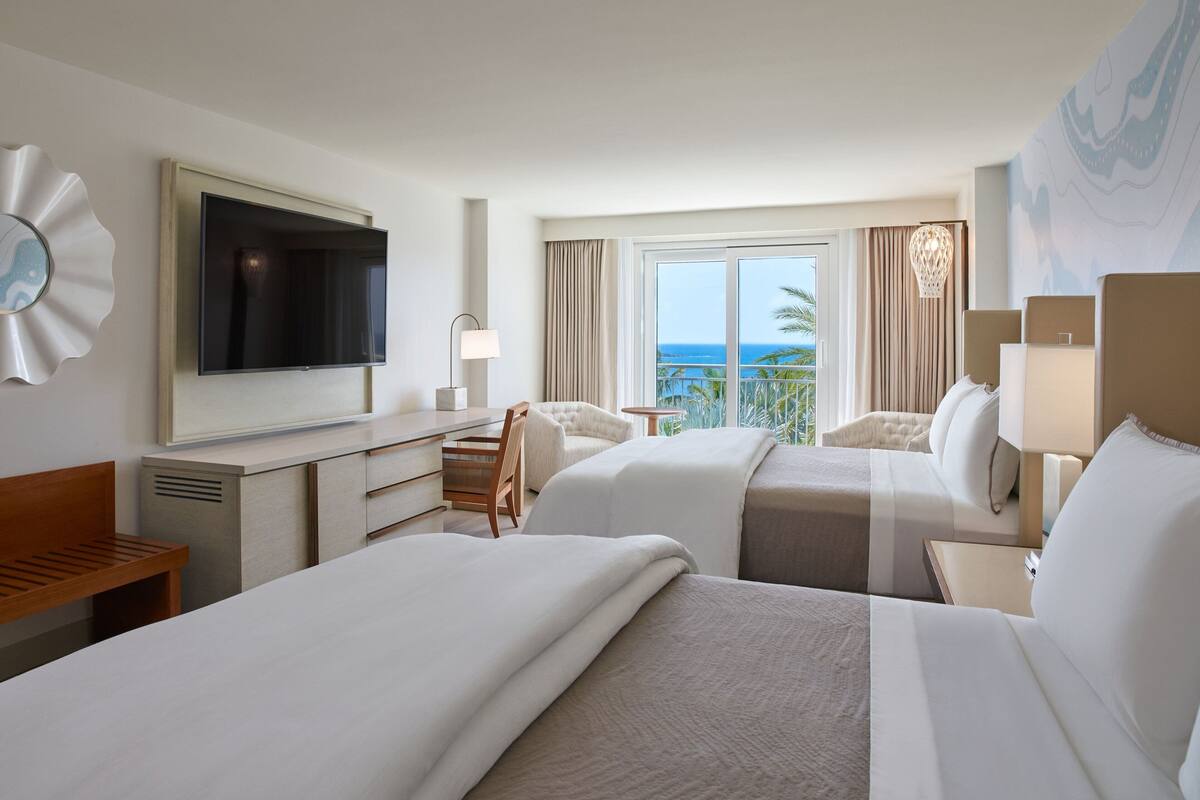 A hotel room with two beds, a TV, a desk, and a view of the ocean.