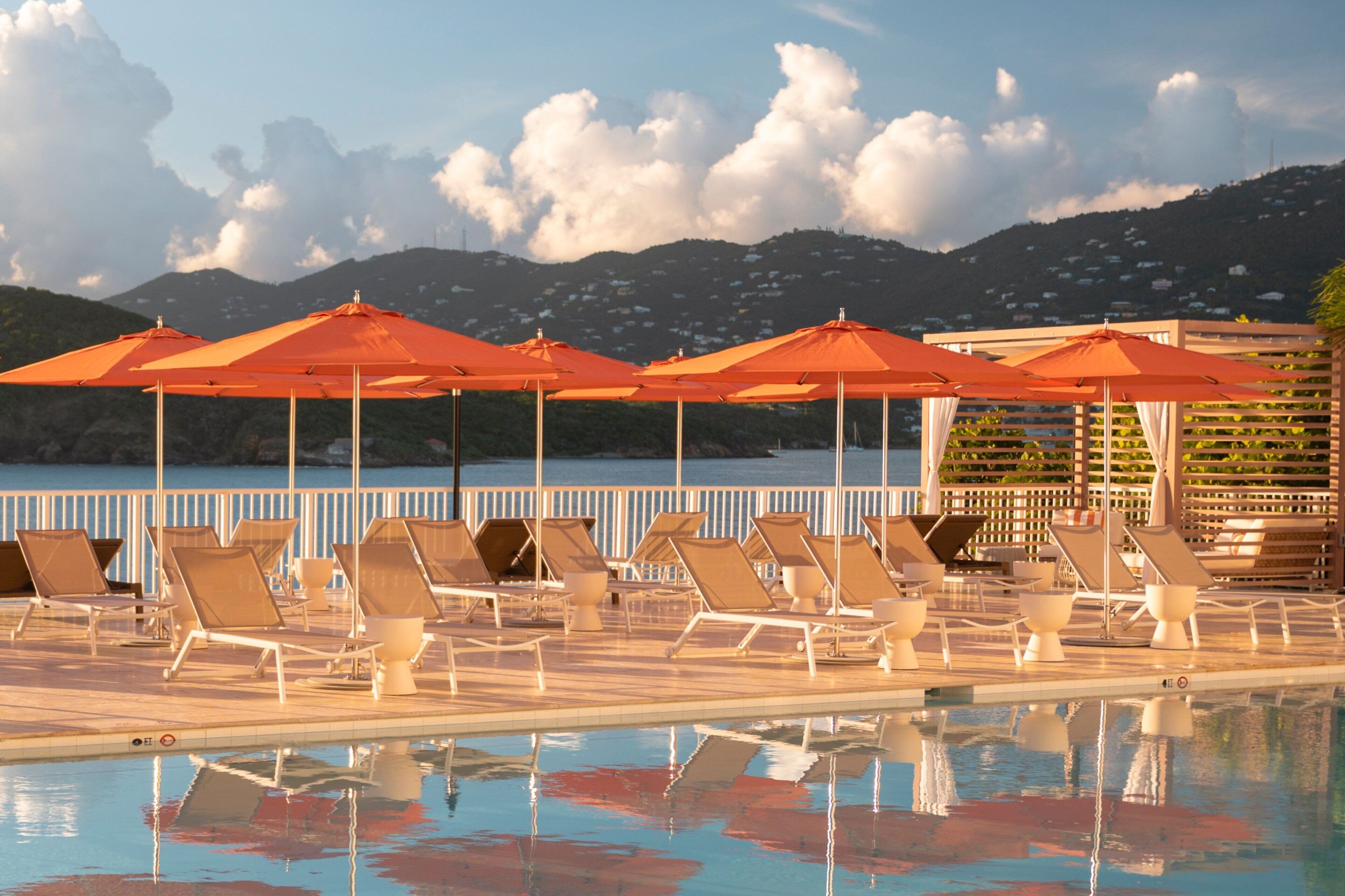Un espace aménagé au bord de la piscine, avec des chaises longues et des parasols orange, offrant une vue dégagée sur l’eau et un paysage montagneux en toile de fond.