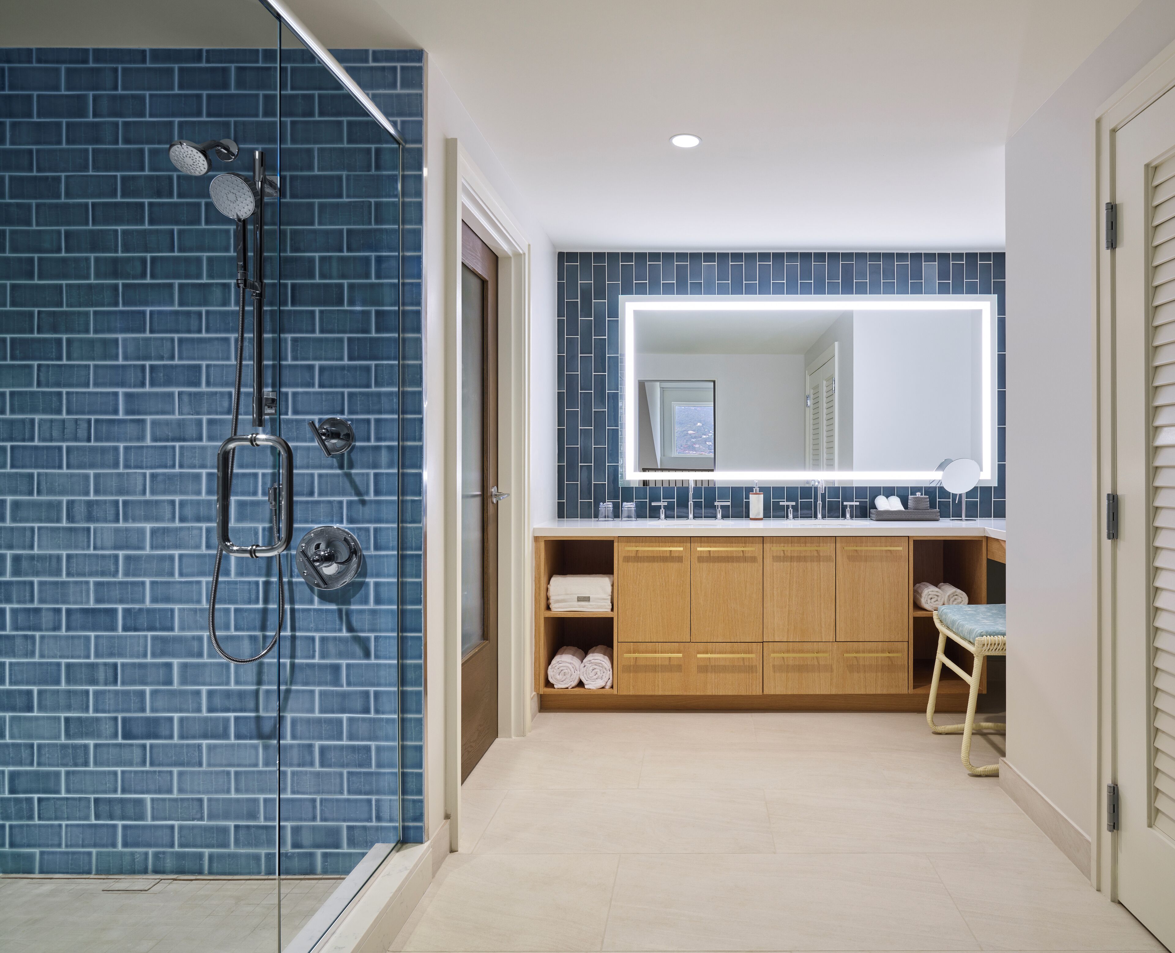 A bathroom with a blue-tiled shower, wooden cabinets, a large mirror, and a towel rack.
