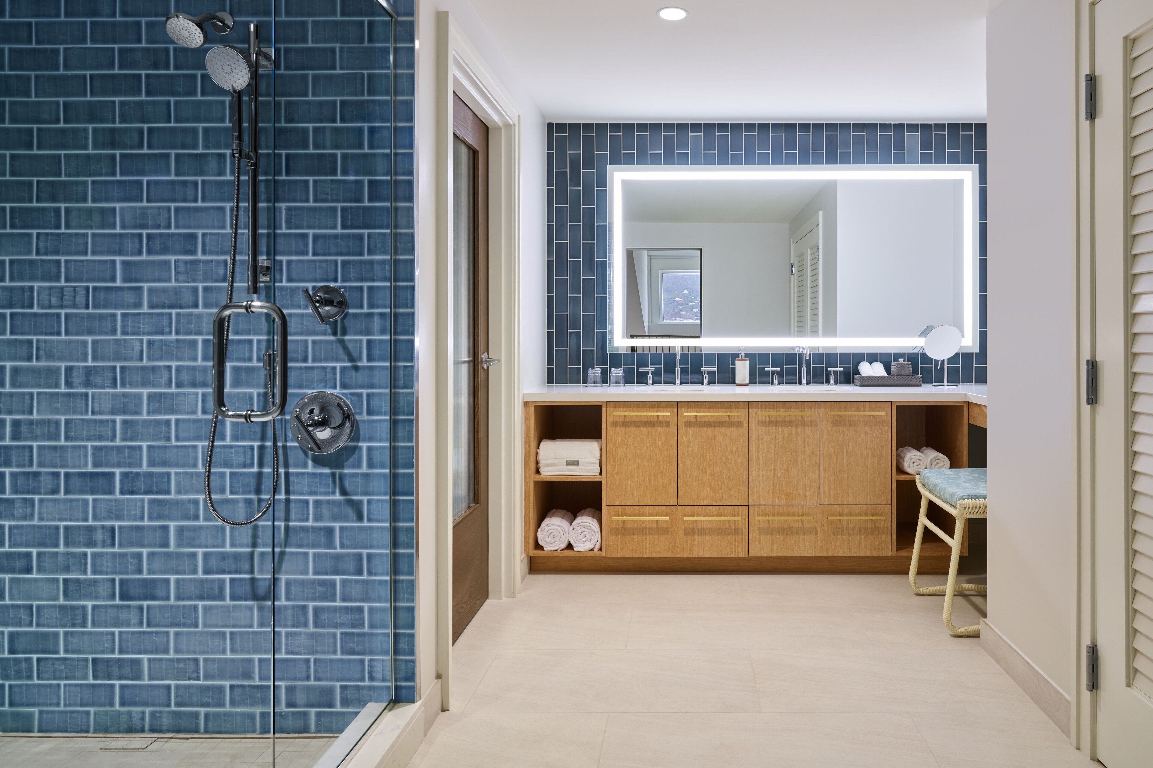 Un baño con pared de azulejos azules, tocador de madera con espejo y ducha con puerta de vidrio.