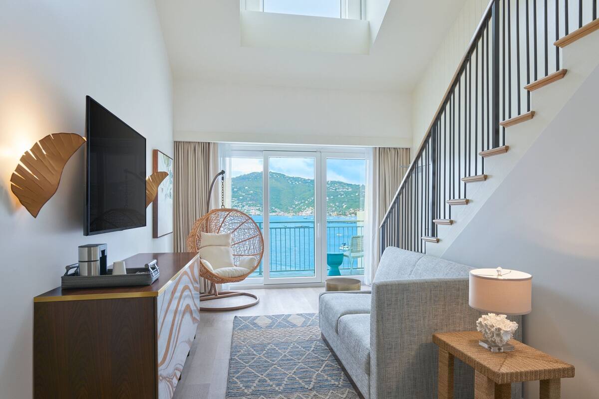 A modern living room with a sofa, a hanging chair, a TV, a small table, and a lamp. A staircase leads to a balcony with a view of the sea.