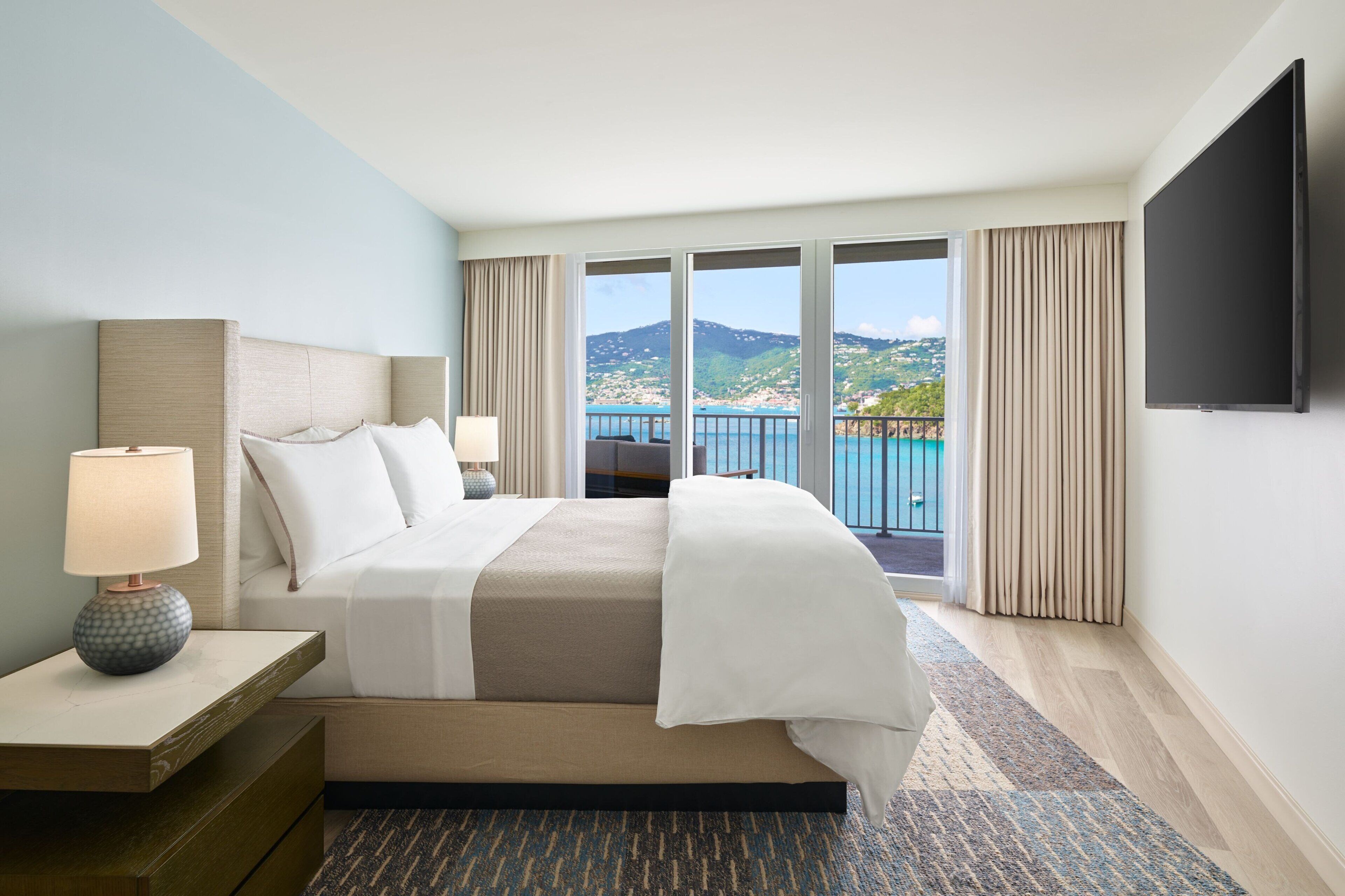 A hotel room with a large bed, a TV, a nightstand, and a view of a coastal landscape.