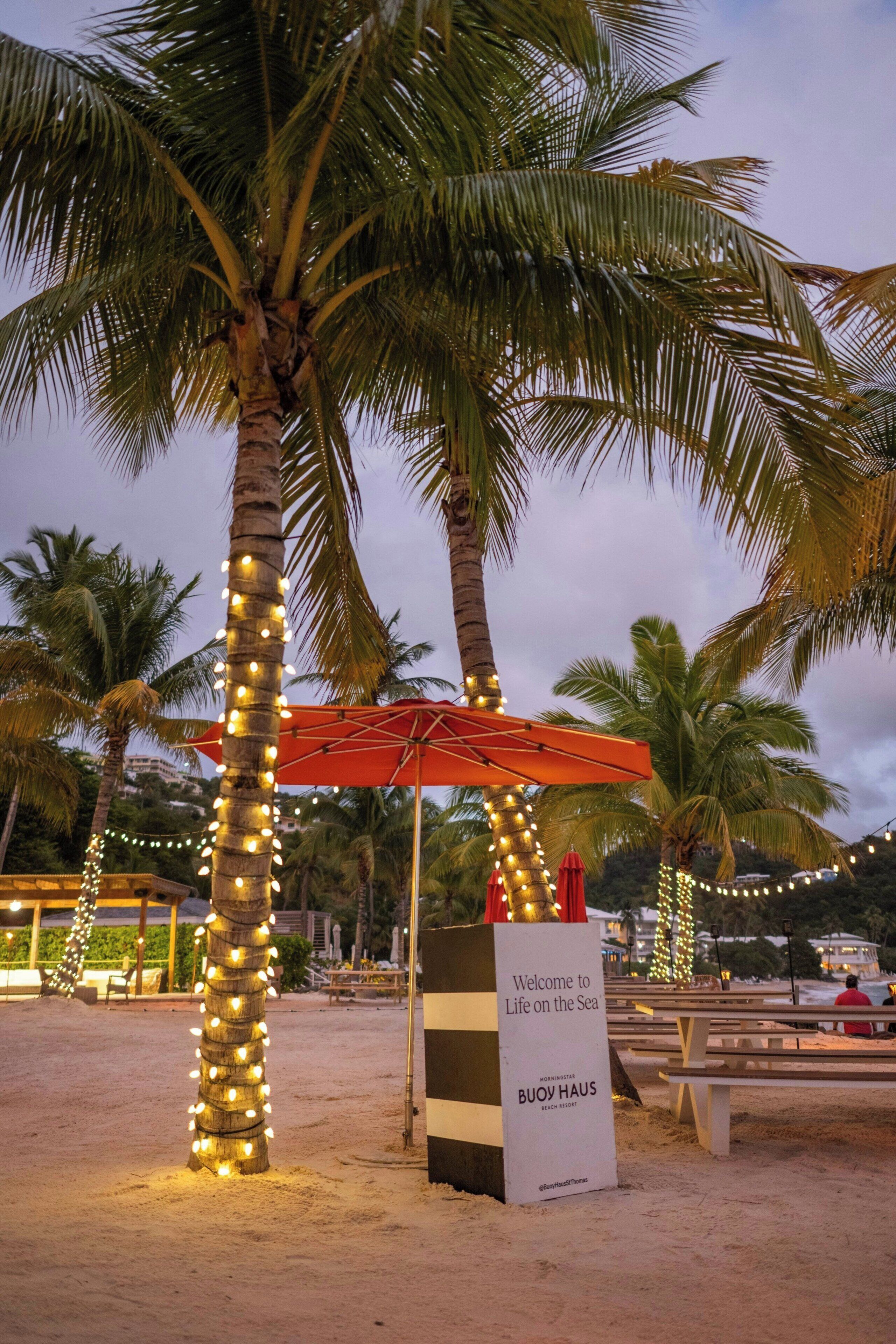 Una zona de playa con palmeras, una sombrilla roja y un cartel que da la bienvenida a visitantes a Life on the Sea.