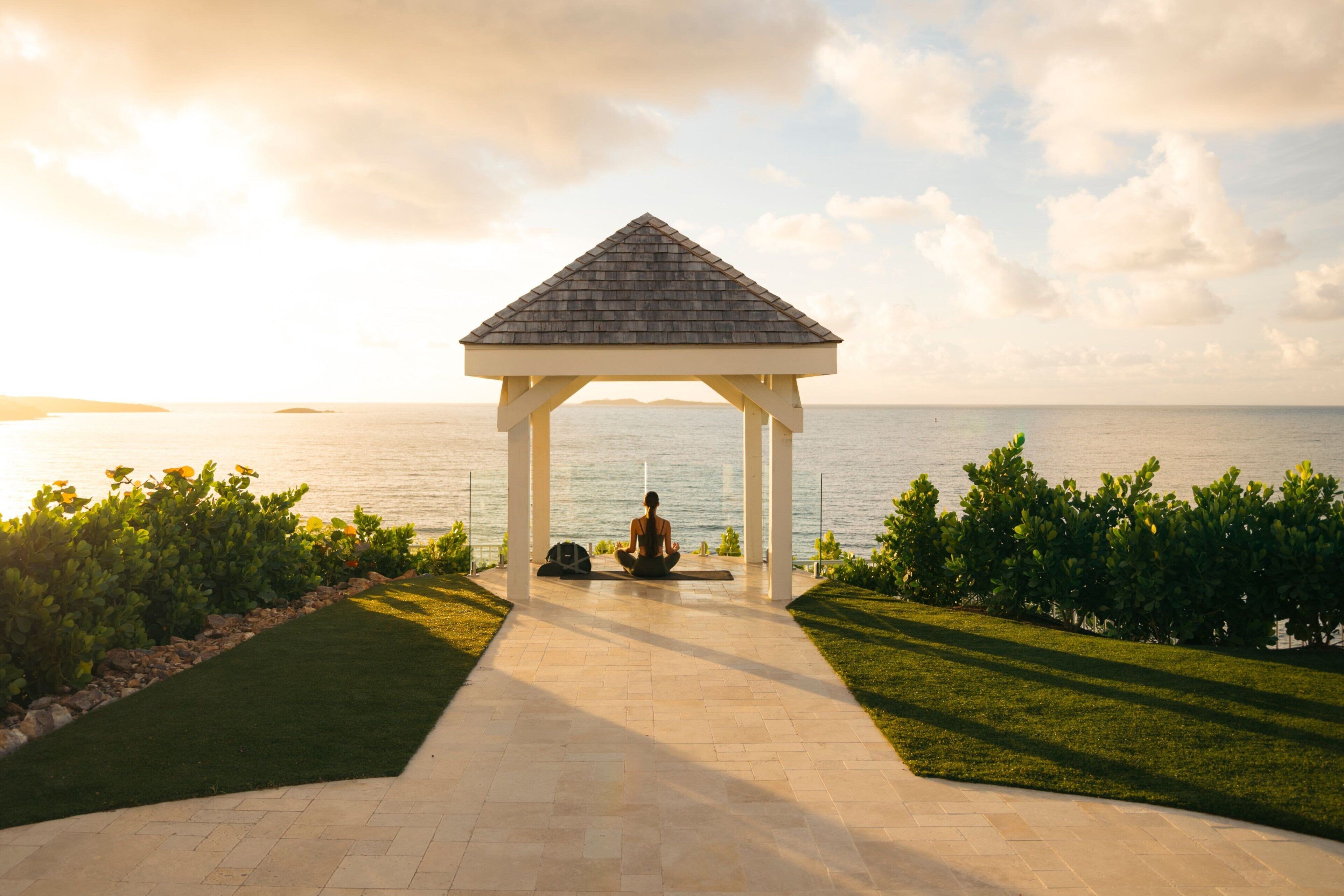 A gazebo with a person meditating, a paved pathway, and a view of the sea.