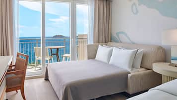 A hotel room with a bed, a desk, and a chair. There is a balcony with a view of the sea and a distant island.