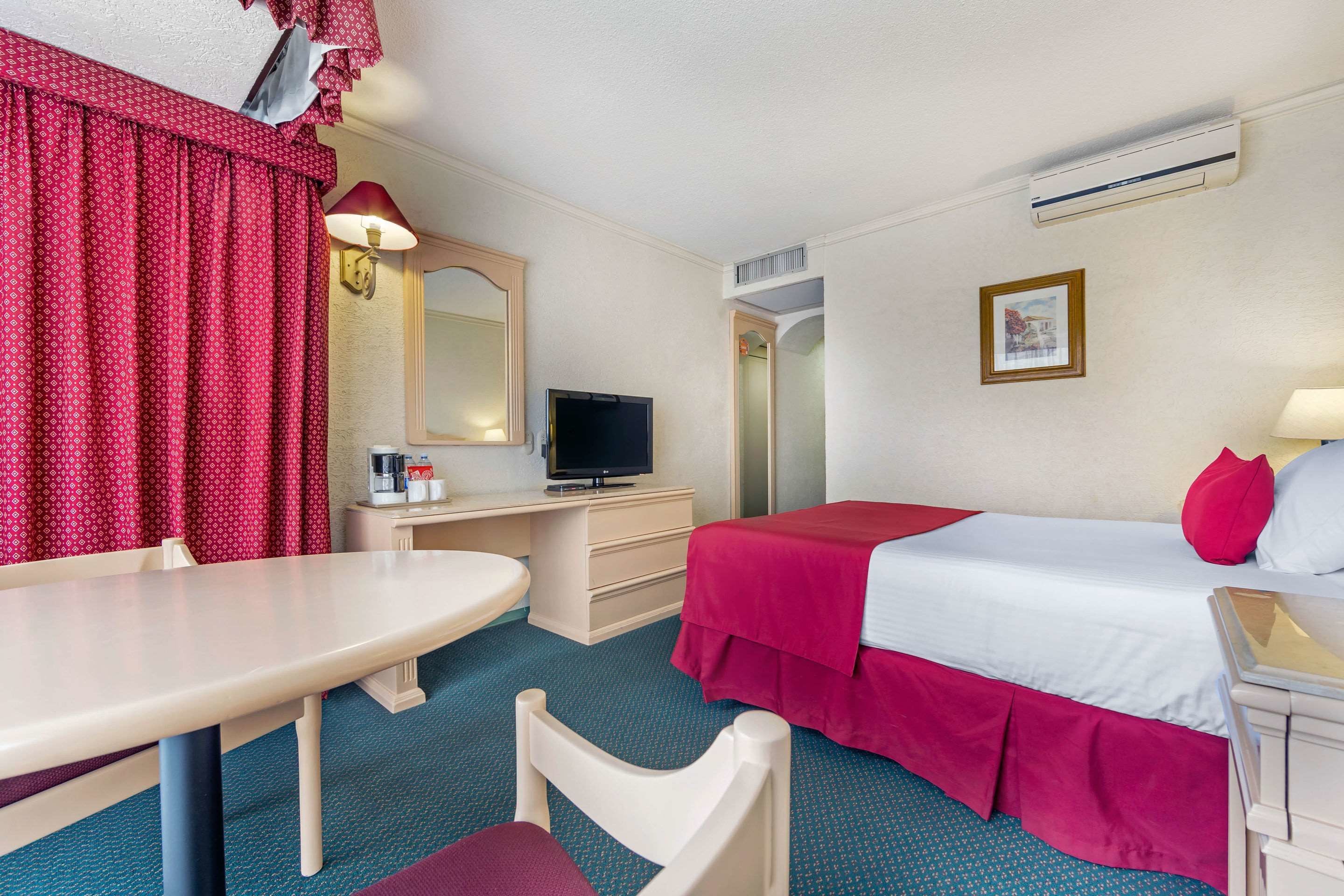 Une chambre d’hôtel comprenant un lit, un bureau avec une télévision, une chaise, une table et une fenêtre avec des rideaux.
