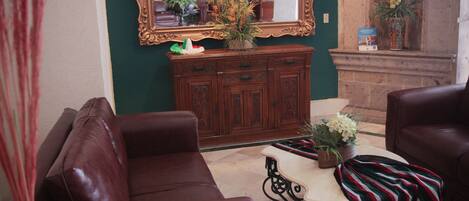 Un salón con un sofá de cuero marrón, una mesita con mantel a rayas, un jarrón decorativo y un espejo grande en la pared.