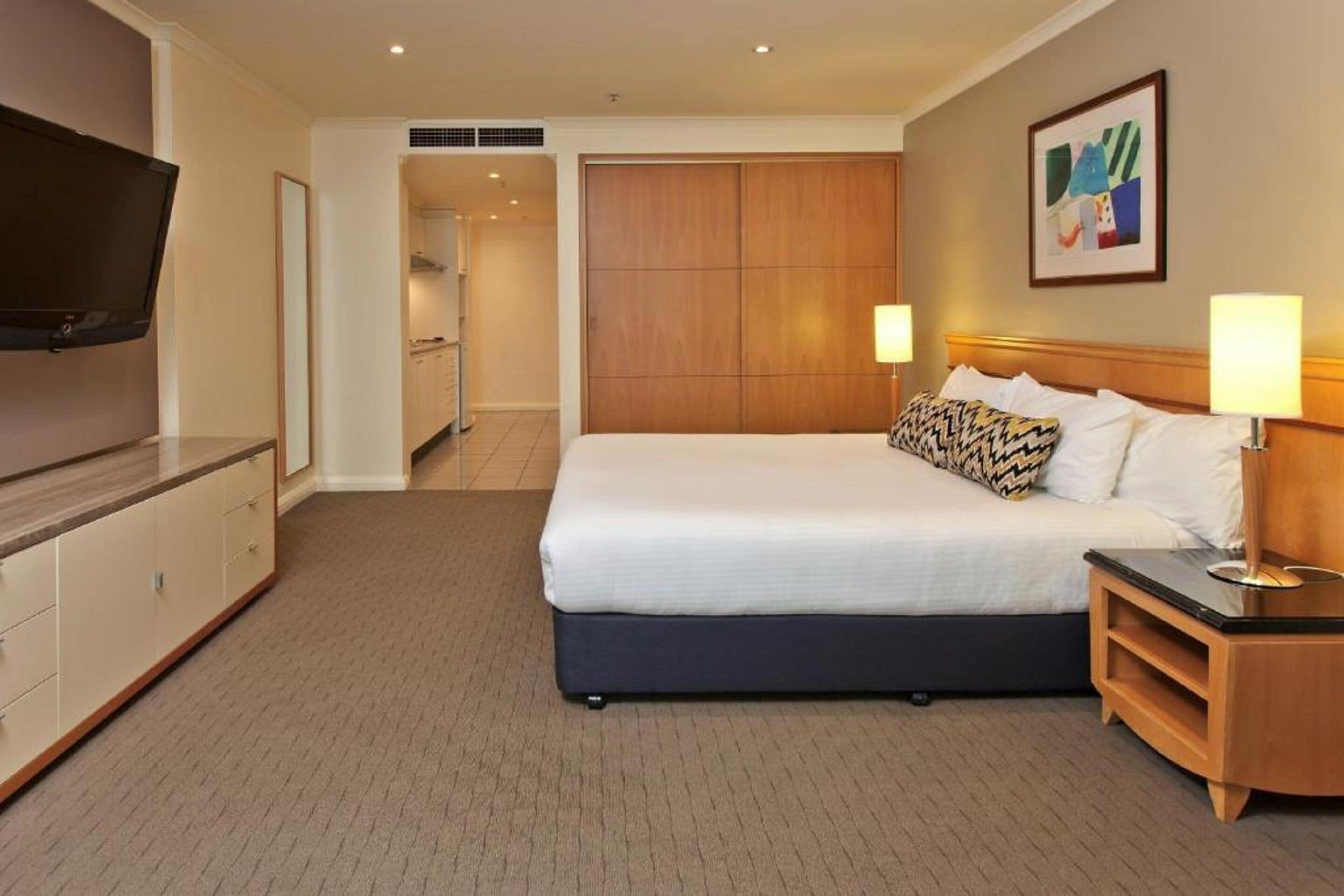 Quarto de hotel com cama, luminárias de cabeceira, televisão, guarda-roupa e um quadro na parede.