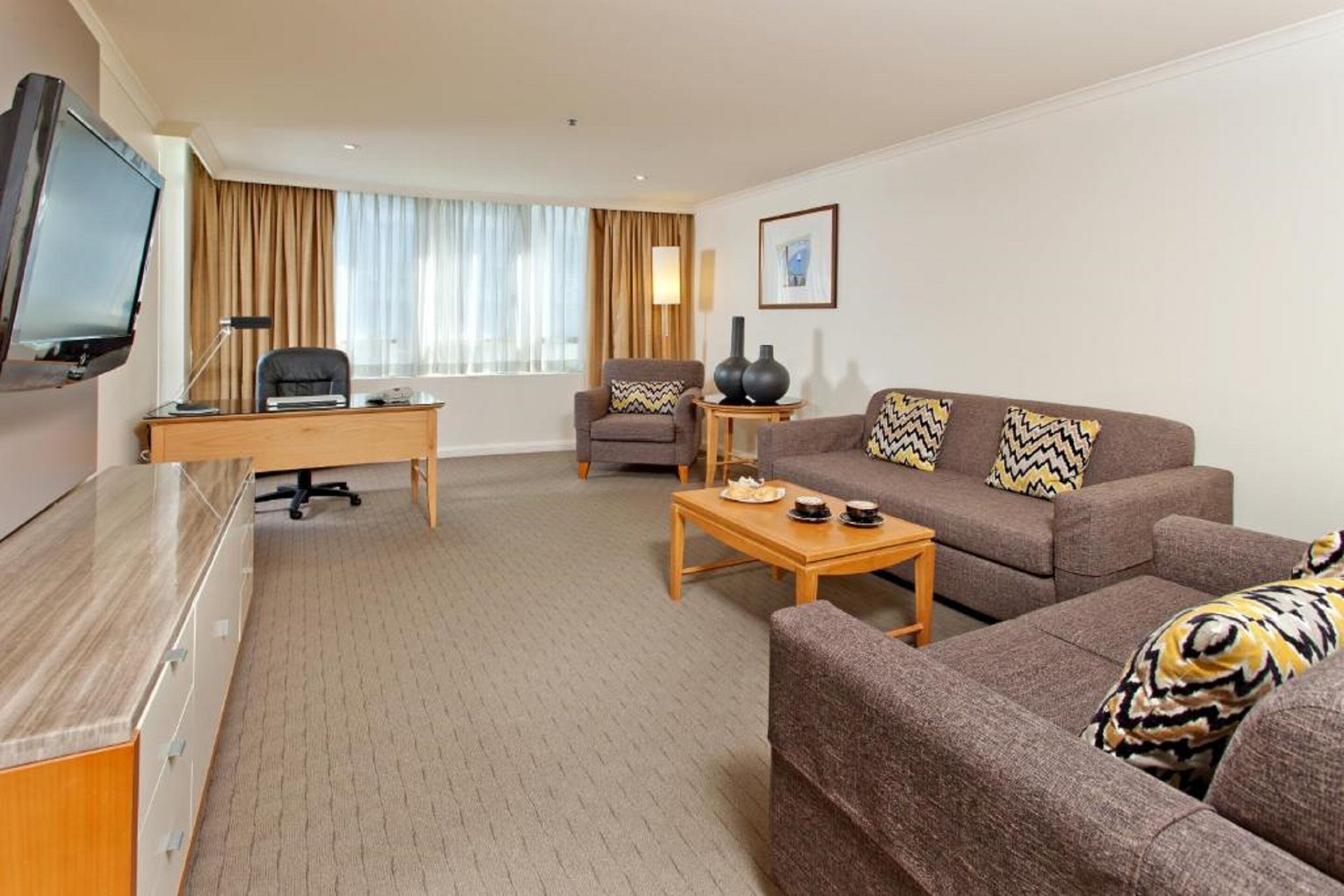 Quarto de hotel com televisão, escrivaninha, sofá, mesa de centro e uma cadeira.