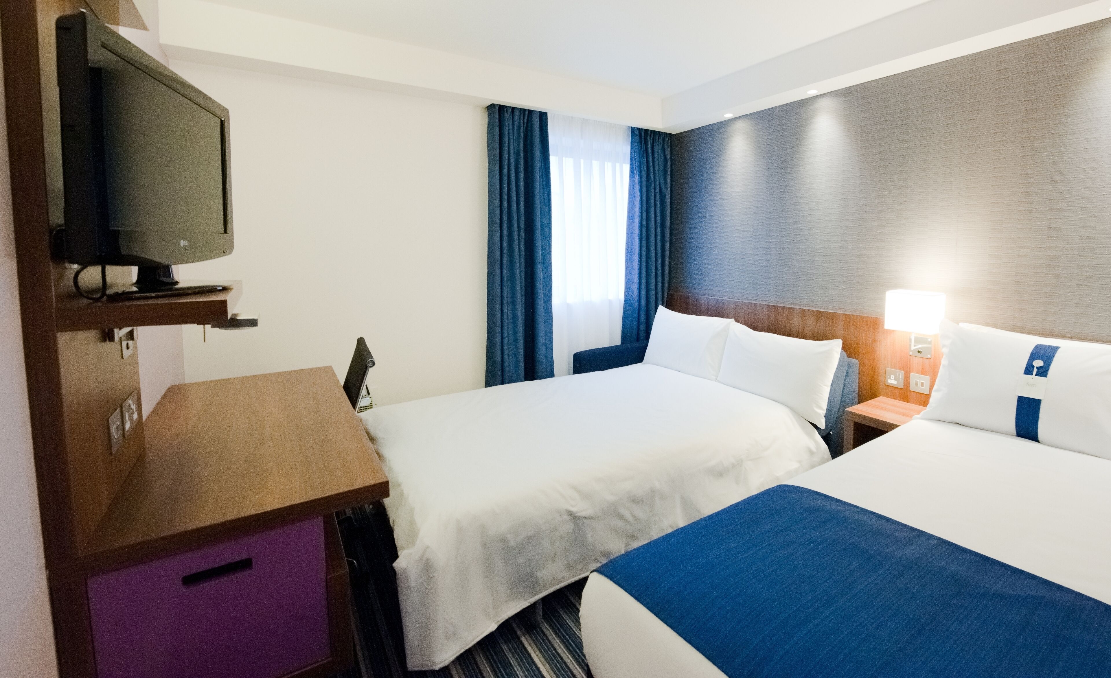 Ein Hotelzimmer mit zwei Betten, einem Schreibtisch, einem Fernseher und einem Fenster mit Vorhängen.