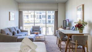 A hotel room with a bed, a sofa, a dining table with chairs, a TV, and a balcony with a view of buildings. - Seasons Botanic Gardens (Melbourne)