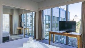 A hotel room with a large bed, a TV on a wooden table, and a view of modern buildings through large windows. - Seasons Botanic Gardens (Melbourne)