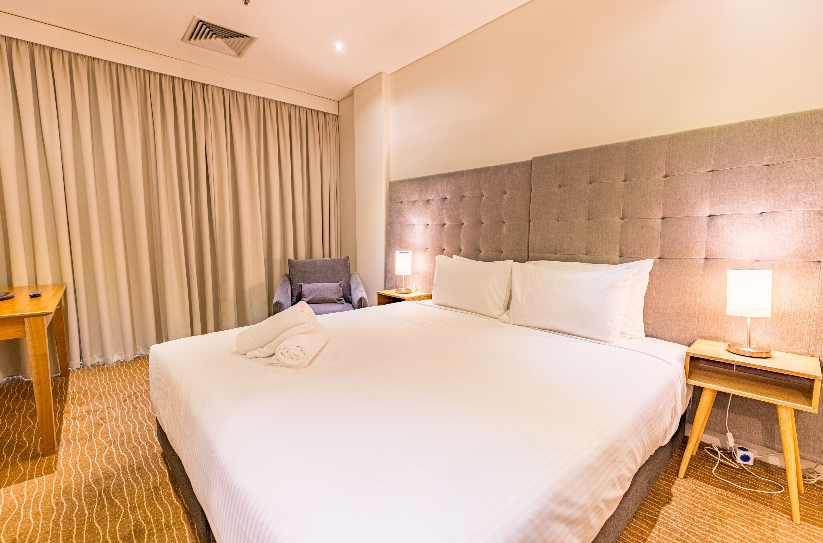 A hotel room with a large bed, a headboard, a desk, a chair, and a lamp.
