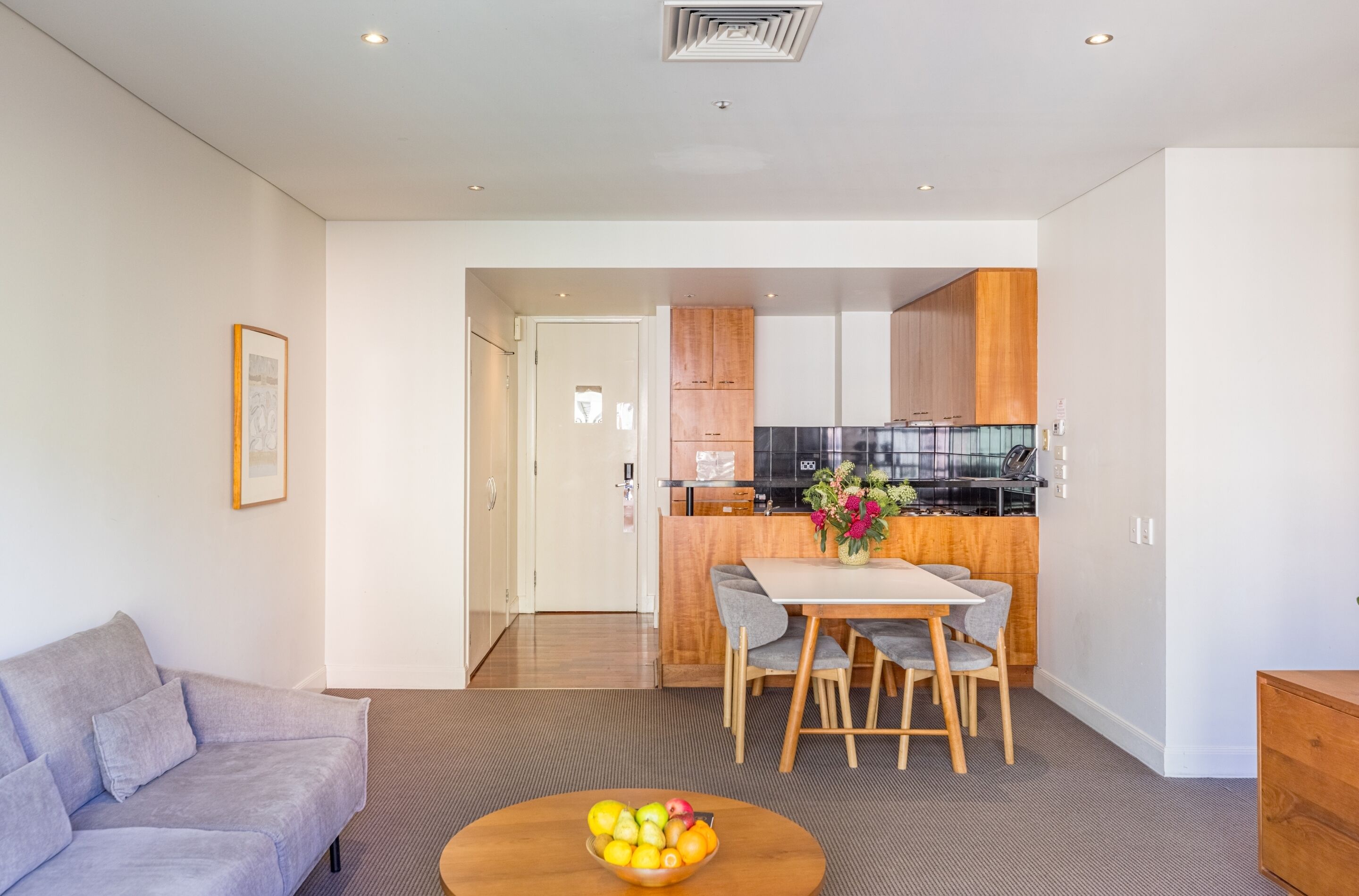 Een moderne woonkamer met een grijze bank, een eettafel met twee stoelen en een fruitschaal. Achtergrond toont een keuken met houten kasten en een zwart aanrecht.