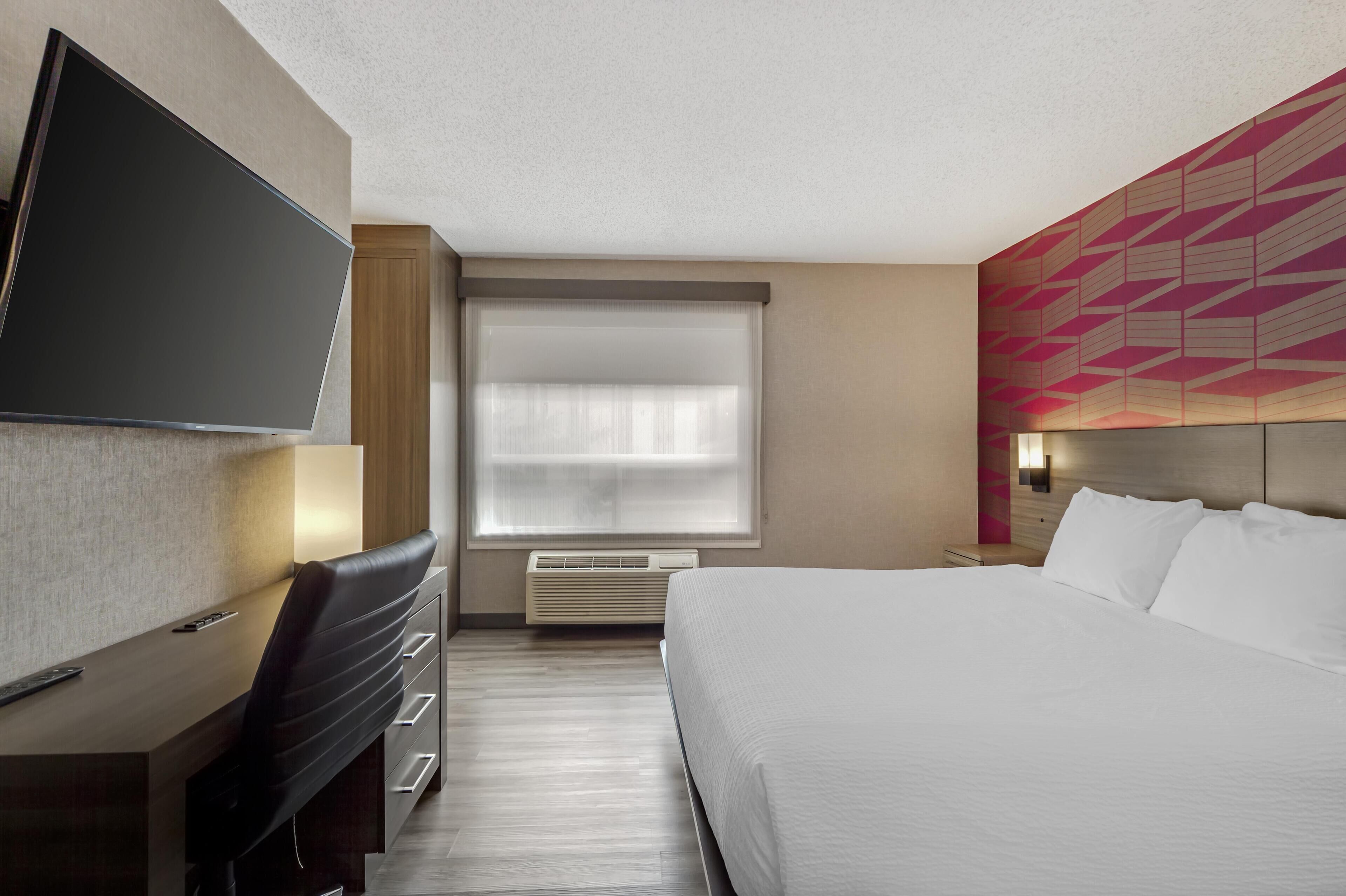 A hotel room with a bed, a desk with a chair, a television, and a window with blinds.