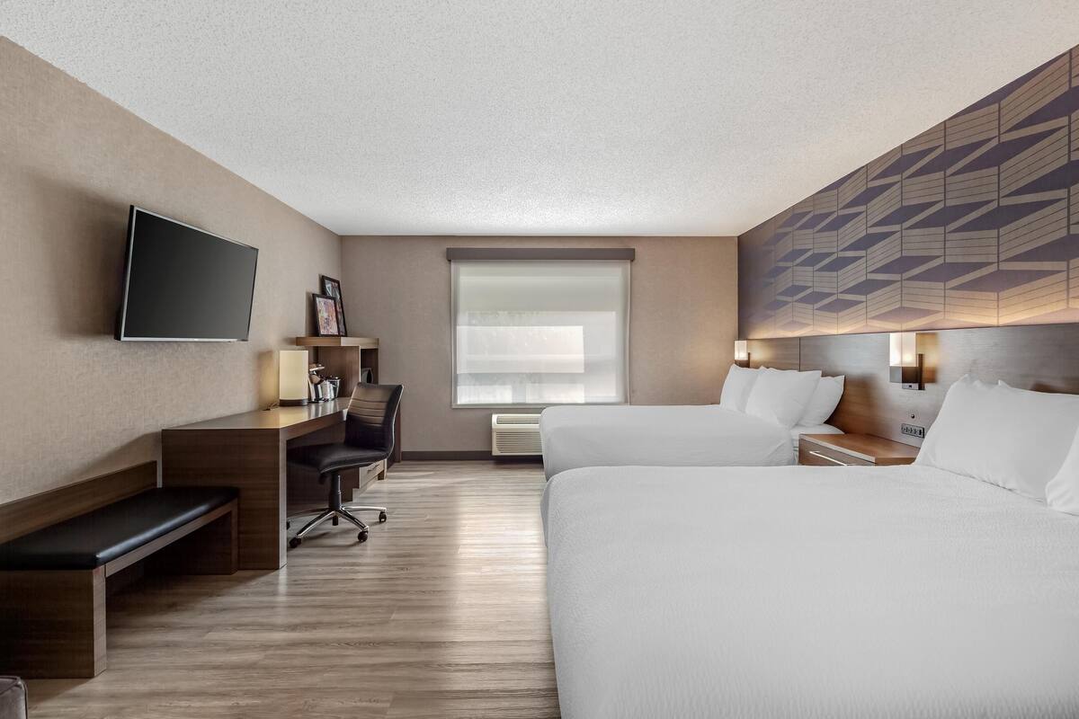 Een hotelkamer met twee bedden, een bureau met stoel, een bankje, een flatscreen televisie en een raam met zonwering.