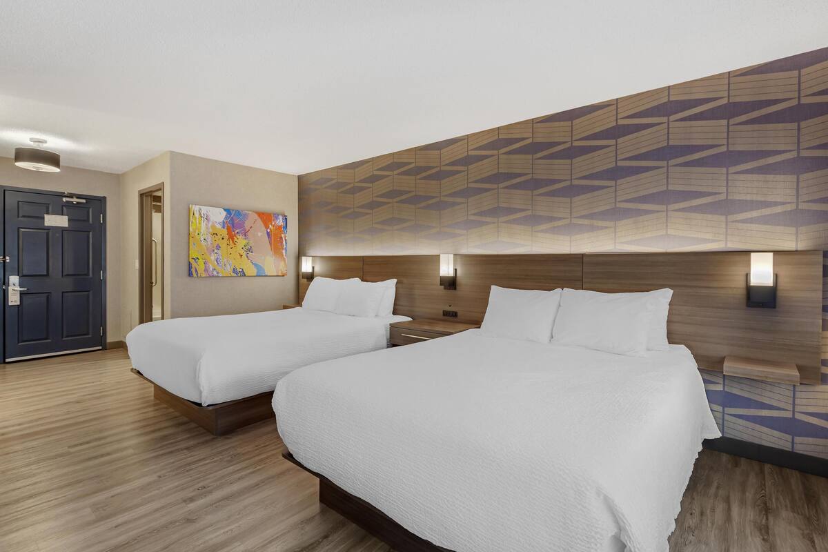 Een hotelkamer met twee bedden, een houten hoofdbord, een kleurrijk abstract schilderij en een deur.