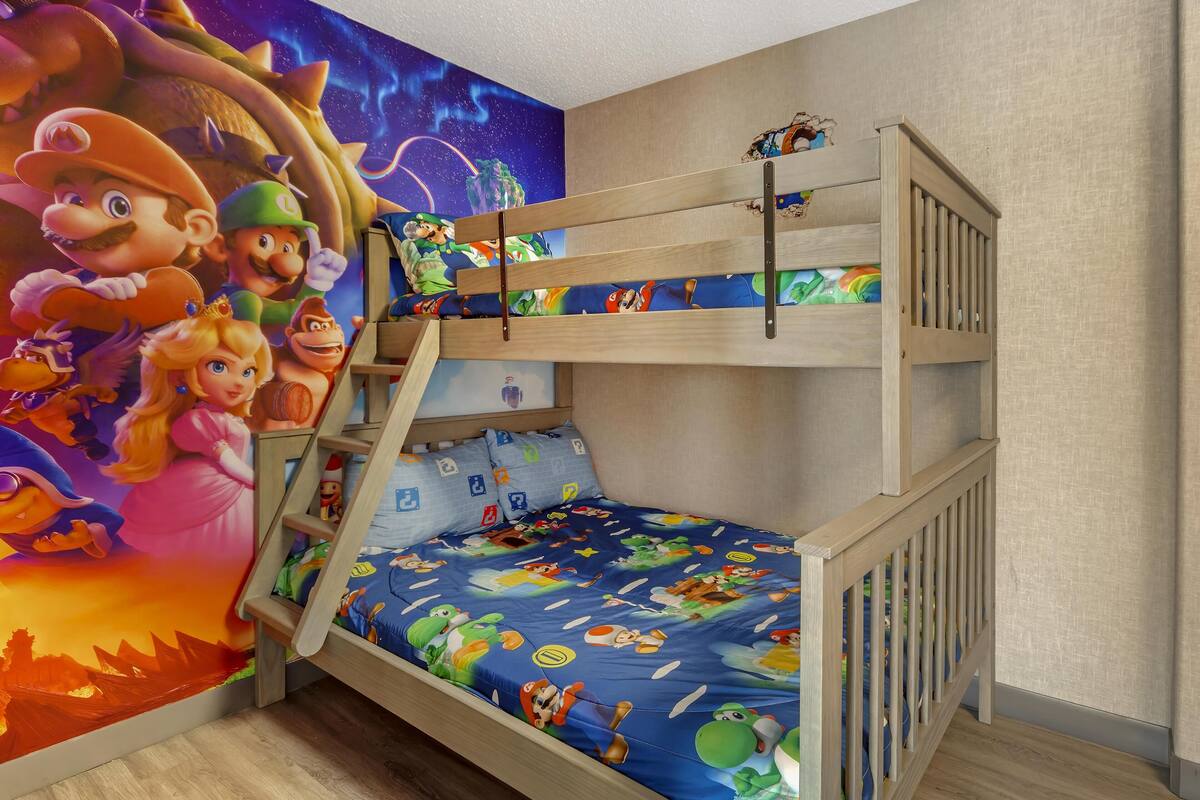 Een stapelbed met een muurschildering van Mario, een houten ladder en een blauw matras met tekenfilmfiguren.