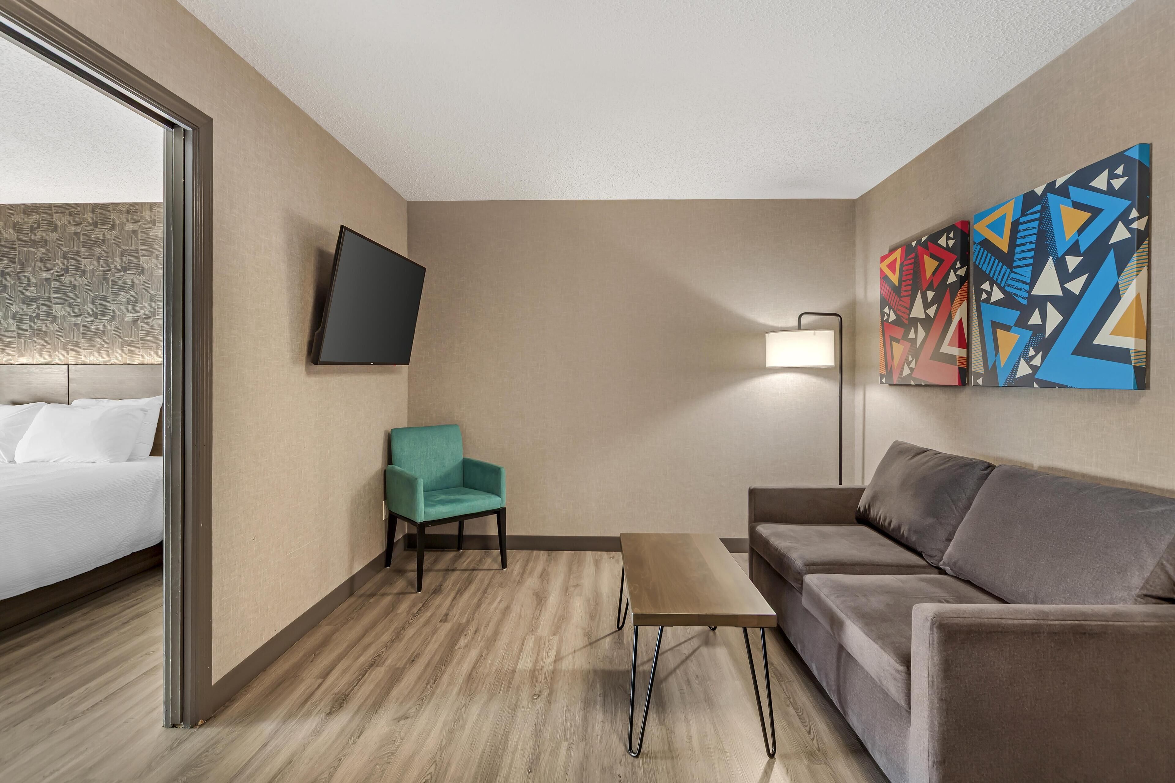 A hotel room with a bed, a teal armchair, a gray sofa, a small table, and abstract wall art.
