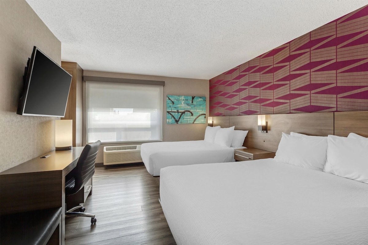 A hotel room with a desk, two beds, a TV, and a window with blinds.