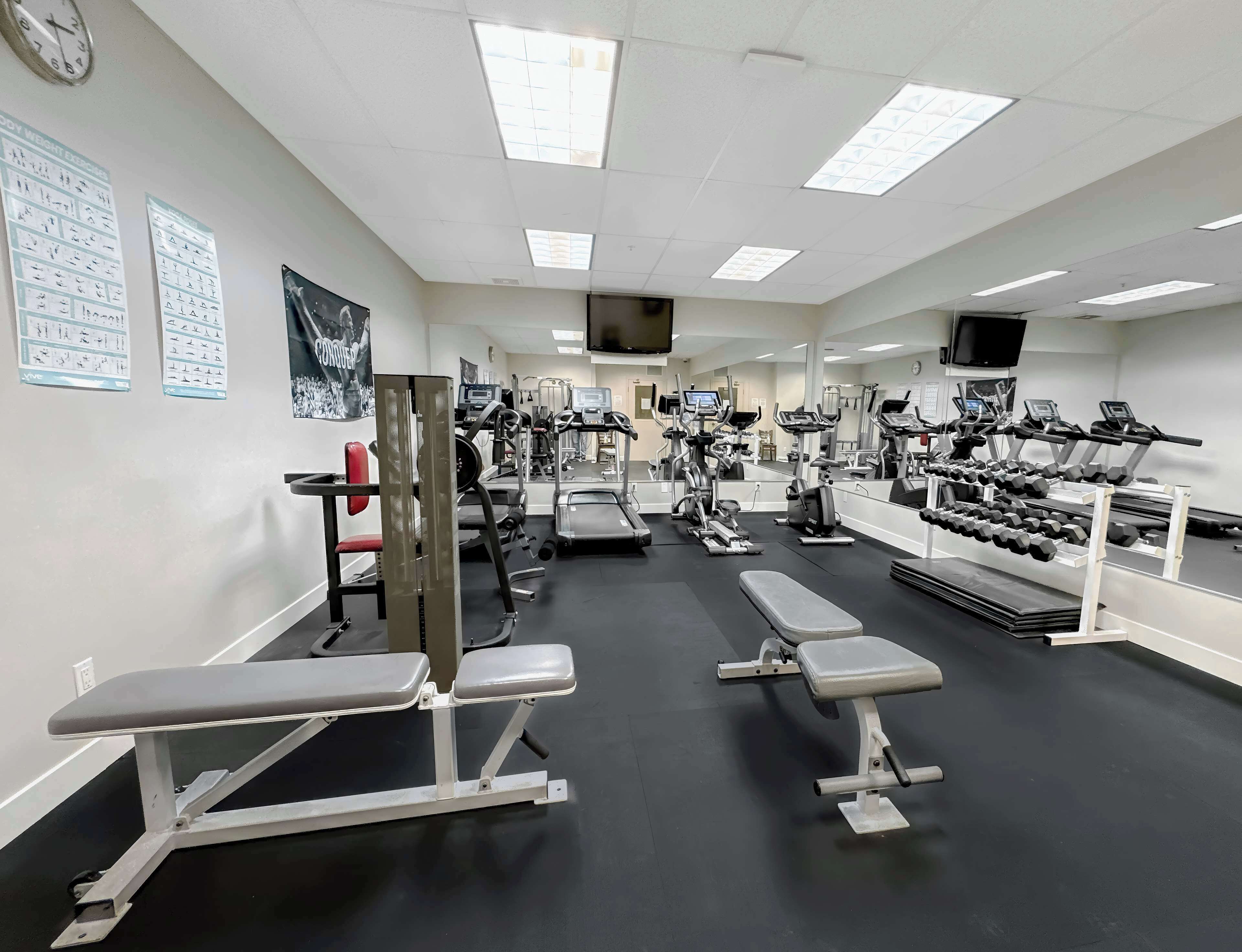 Une salle de sport équipée de divers appareils d’exercice, de miroirs et d’une horloge au mur.