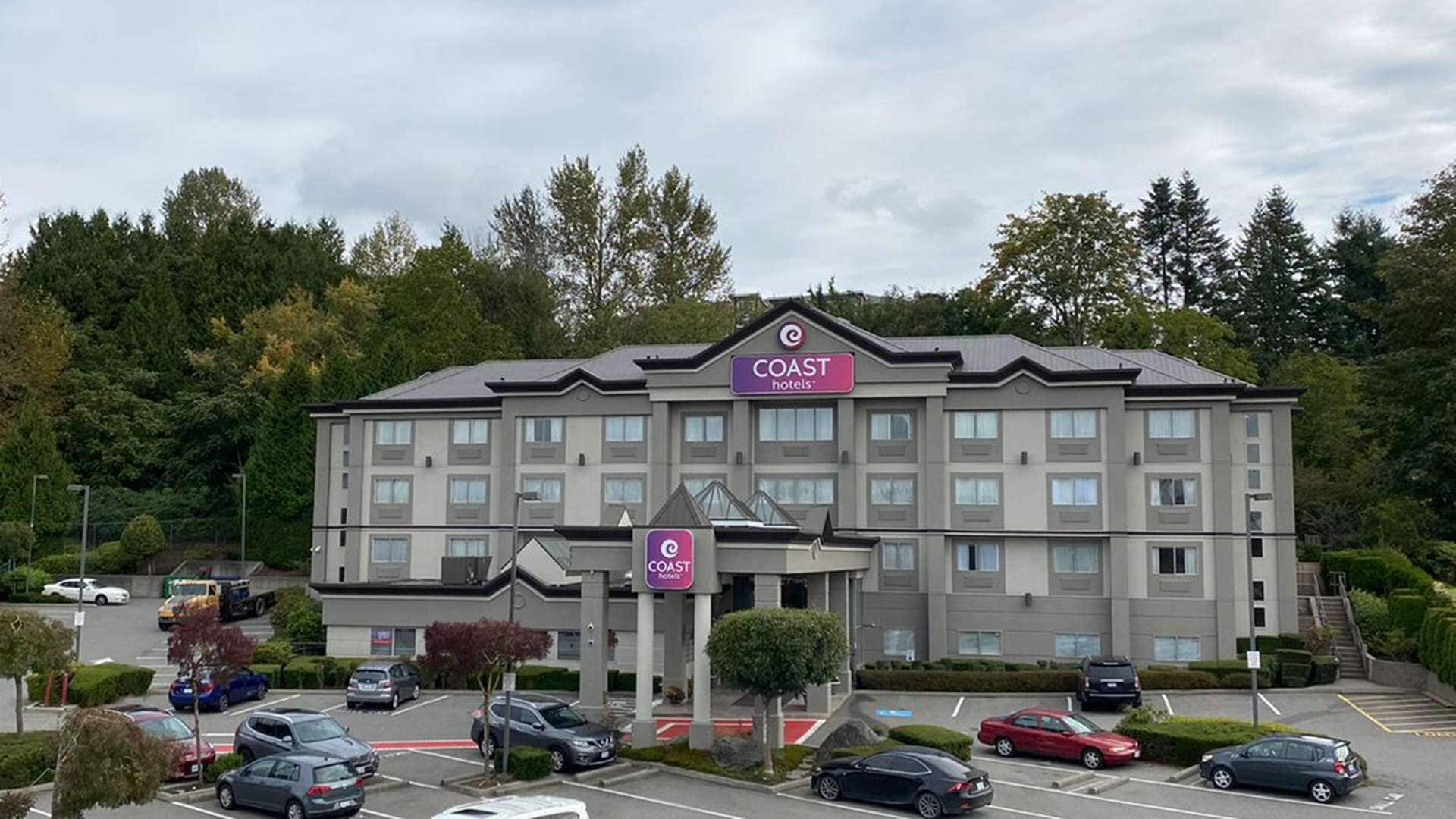 Un hotel de varios pisos con un llamativo letrero de 'Coast Hotels', un estacionamiento con varios coches y áreas verdes circundantes.