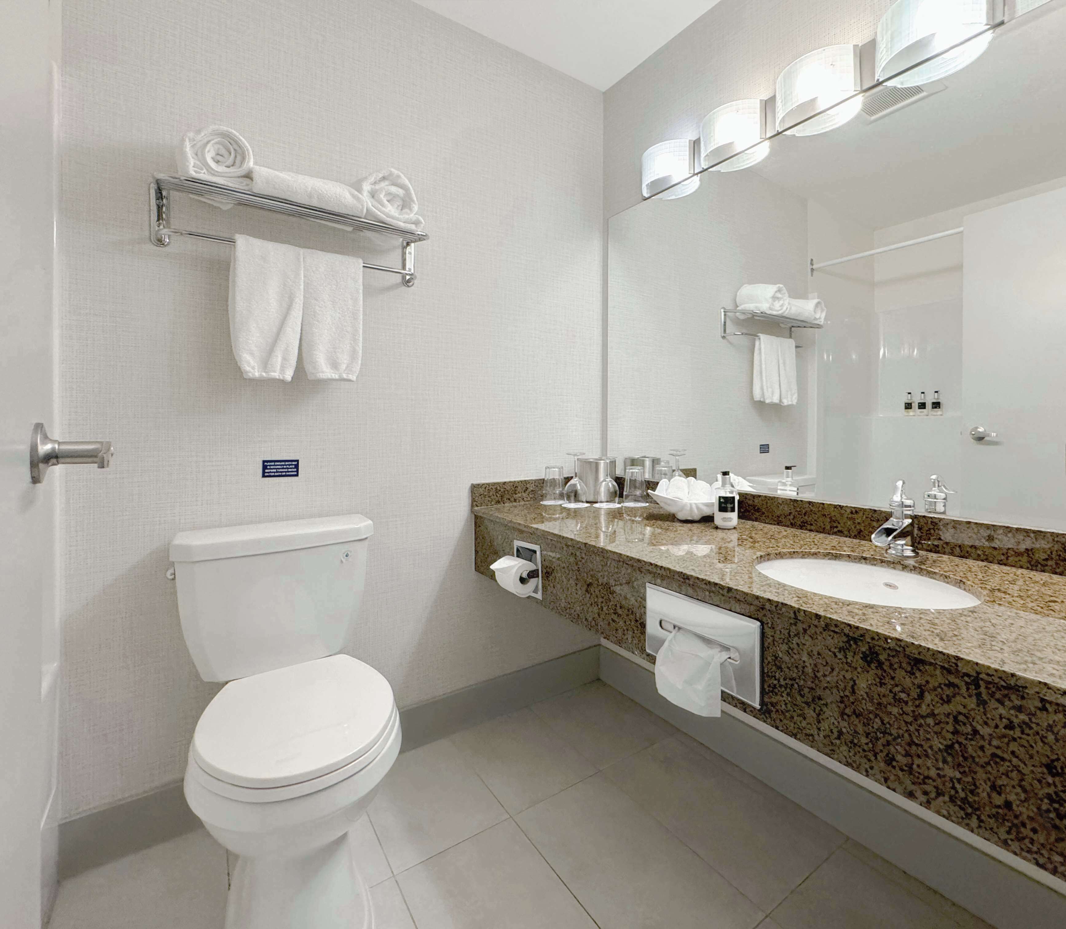 Une salle de bain équipée d’un WC, d’un lavabo, d’un miroir, d’un porte-serviettes et d’un distributeur de serviettes en papier.