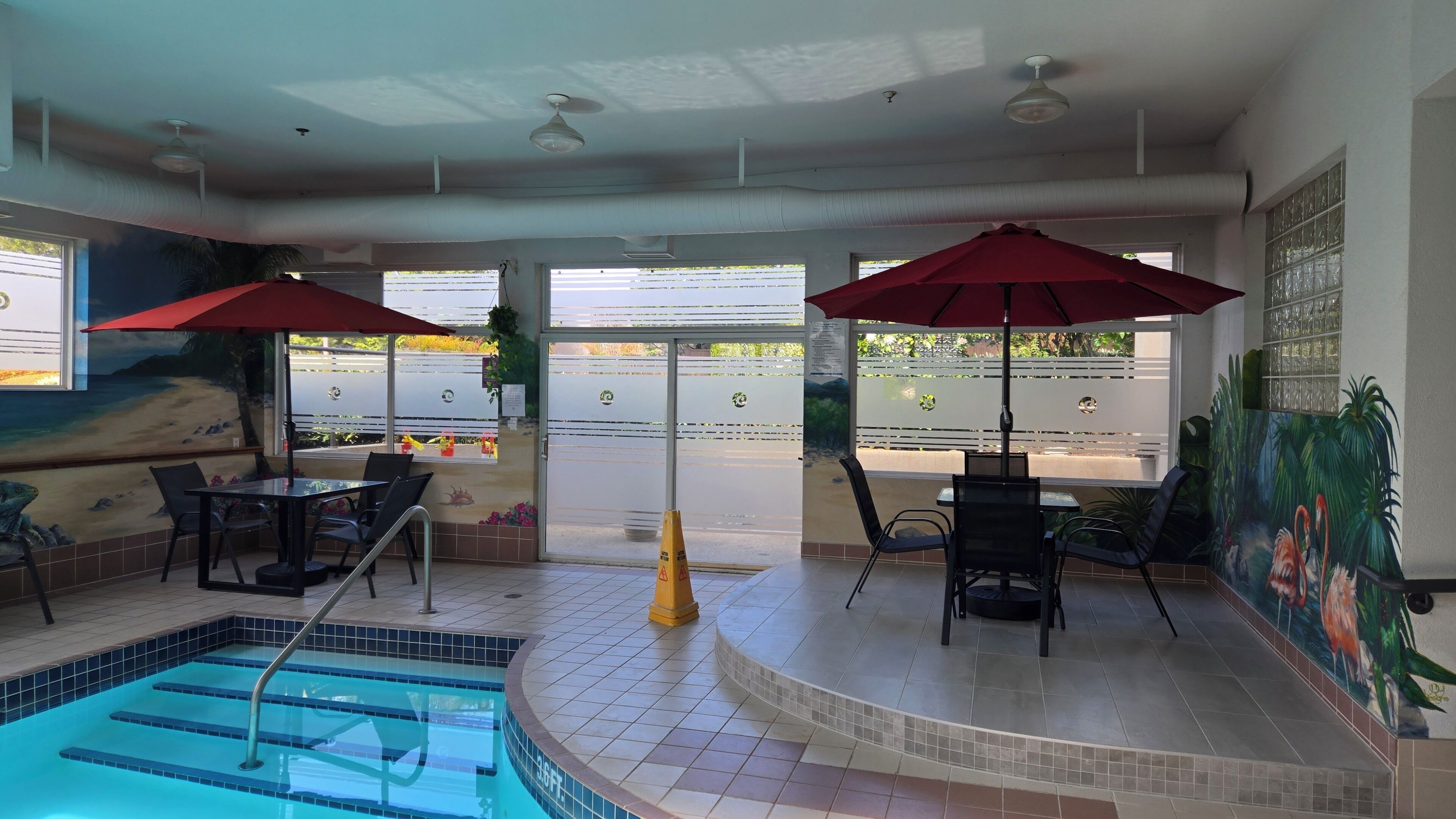 An indoor pool area with a diving board, two red umbrellas, and a mural of a beach scene.
