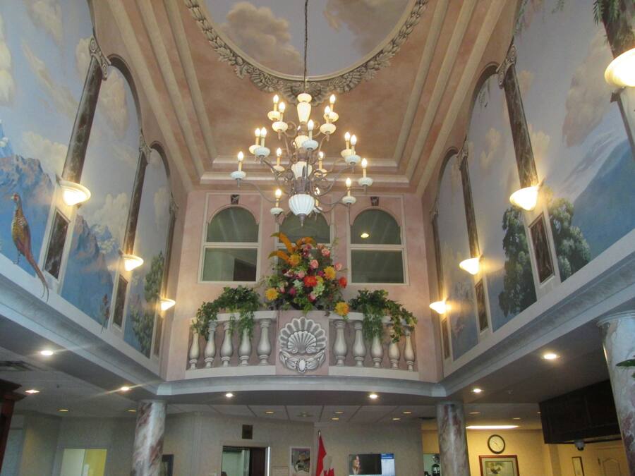 A grand hall with a large chandelier, a balcony with flowers, and a mural of birds on the ceiling.