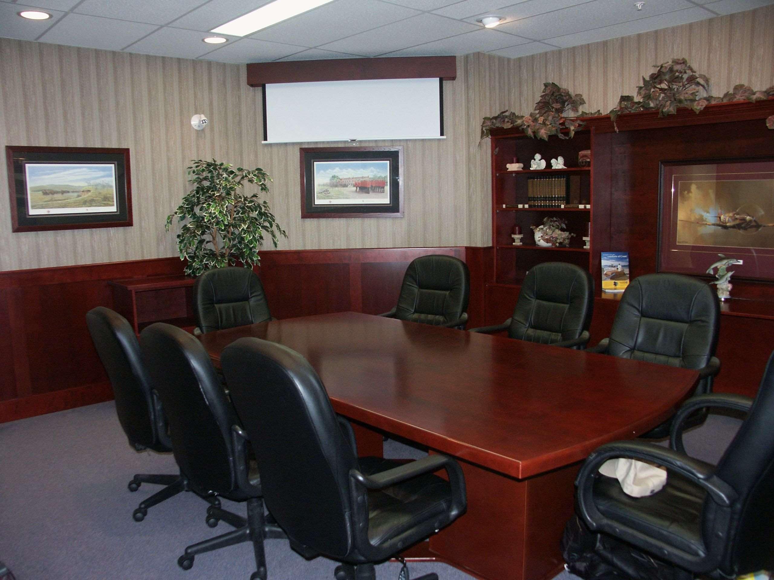A conference room with a large wooden table, six black office chairs, a projector screen, framed pictures, a potted plant, and a decorative shelf with figurines.