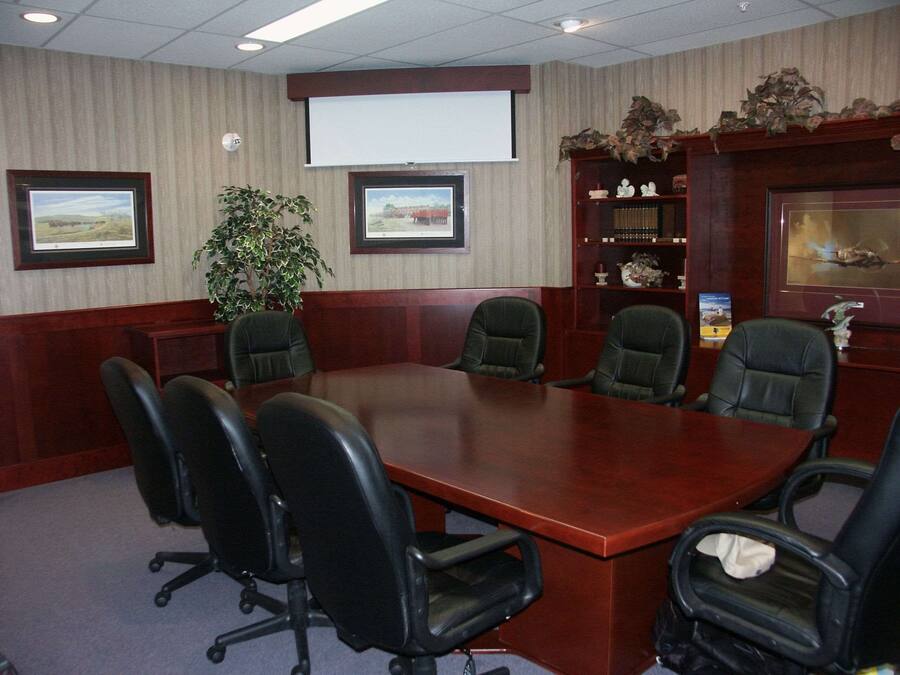 A conference room with a large wooden table, six black office chairs, a projector screen, framed pictures, a potted plant, and a decorative shelf with figurines.