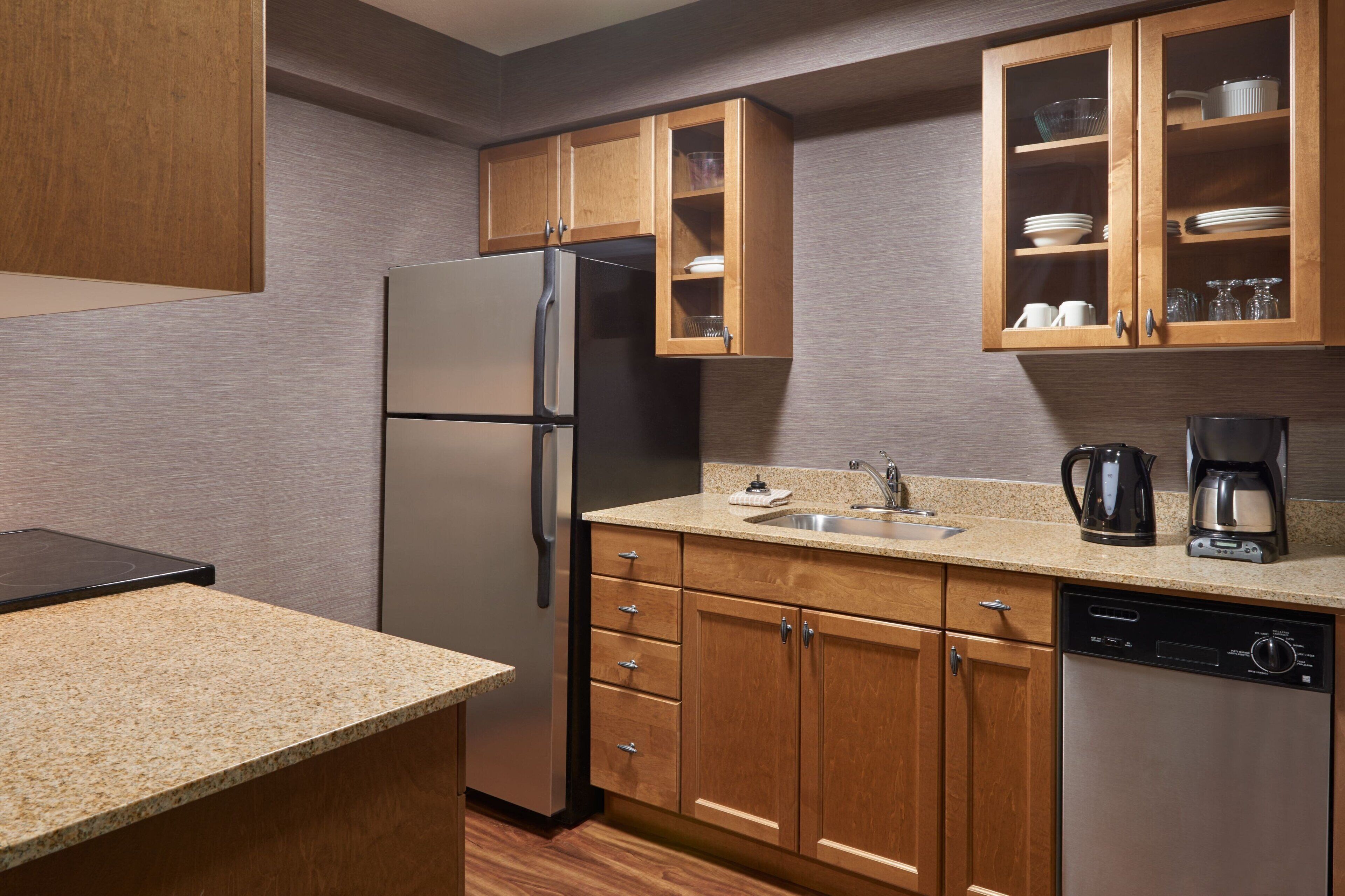 A compact kitchen with wooden cabinets, a stainless steel refrigerator, a dishwasher, and a coffee maker.