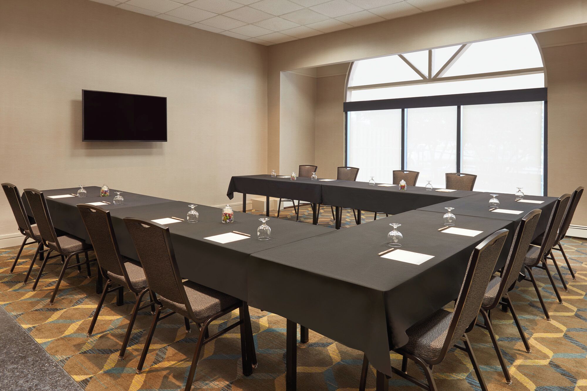 Un salón de conferencias con una mesa larga, sillas, un televisor de pantalla plana y ventanas amplias.