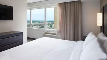 A hotel room with a large bed, a flat-screen TV mounted on the wall, a window with a view of the city, and a desk with drawers.