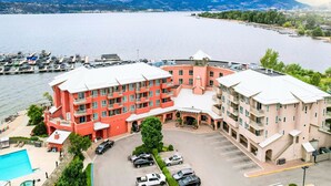 Un hotel de varios pisos con fachada roja y color crema, un estacionamiento y vista a un lago con barcos.