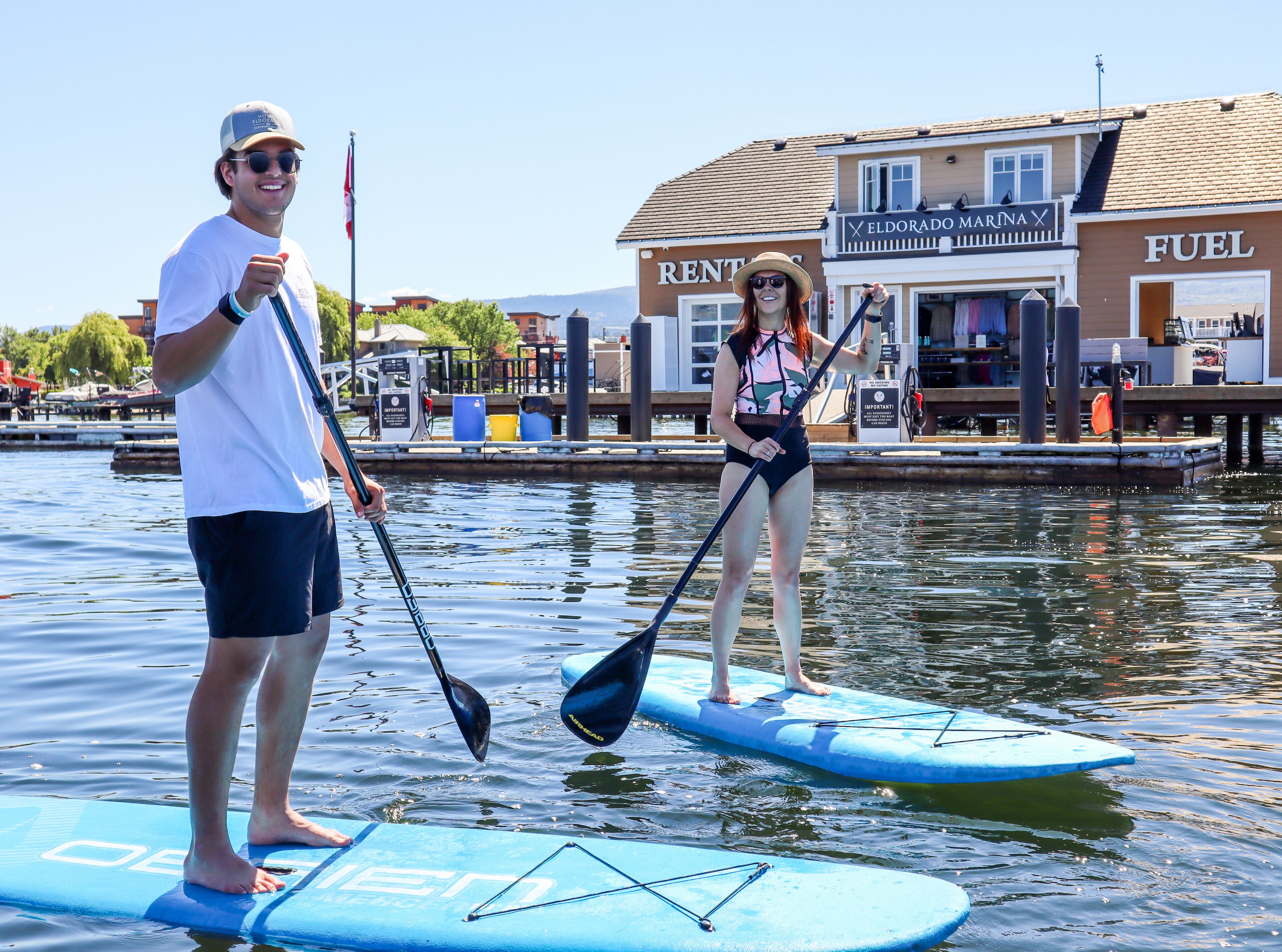 Zwei Personen stehen auf Paddleboards auf ruhigem Wasser, im Hintergrund sind Gebäude zu sehen, darunter ein Schild mit der Aufschrift 'RENTALS'.