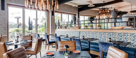 En modern restaurang med lädersitsar, en bardisk med utsikt och ett stort utbud av växter och dekorativa föremål.