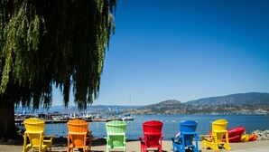 Una hilera de coloridas sillas Adirondack alineadas en una playa arenosa, con vistas a una masa de agua tranquila con barcos y colinas al fondo.