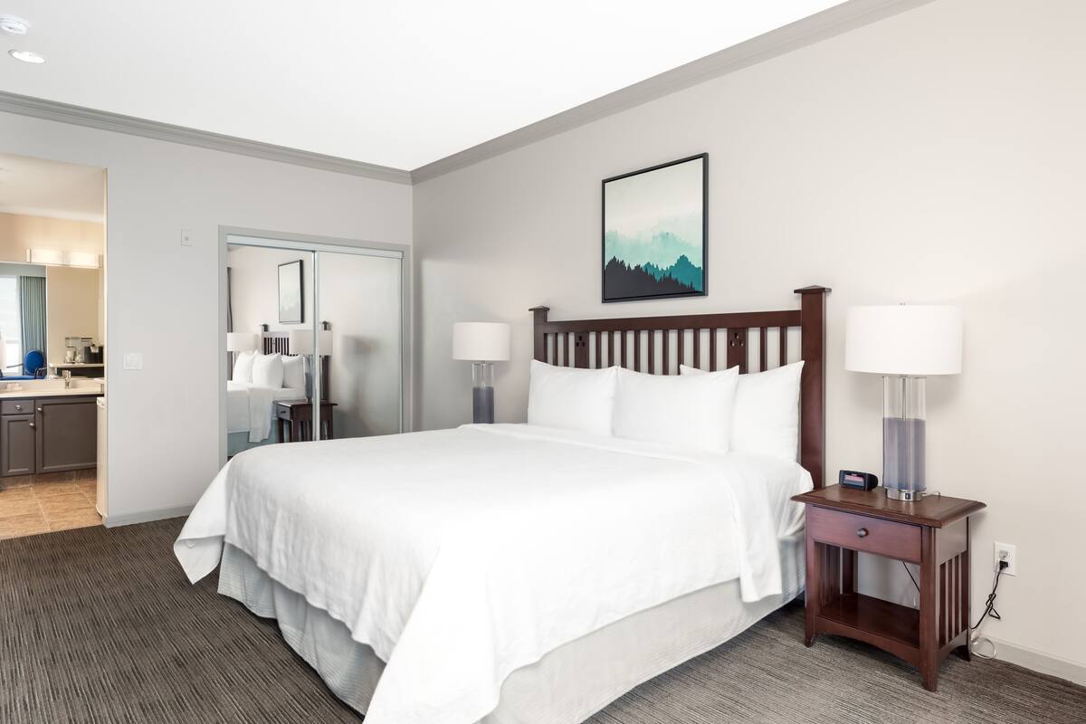 Una habitación de hotel con una cama grande, lámparas de noche, una mesita de noche, un cuadro en la pared y un baño visible a través de una puerta abierta.