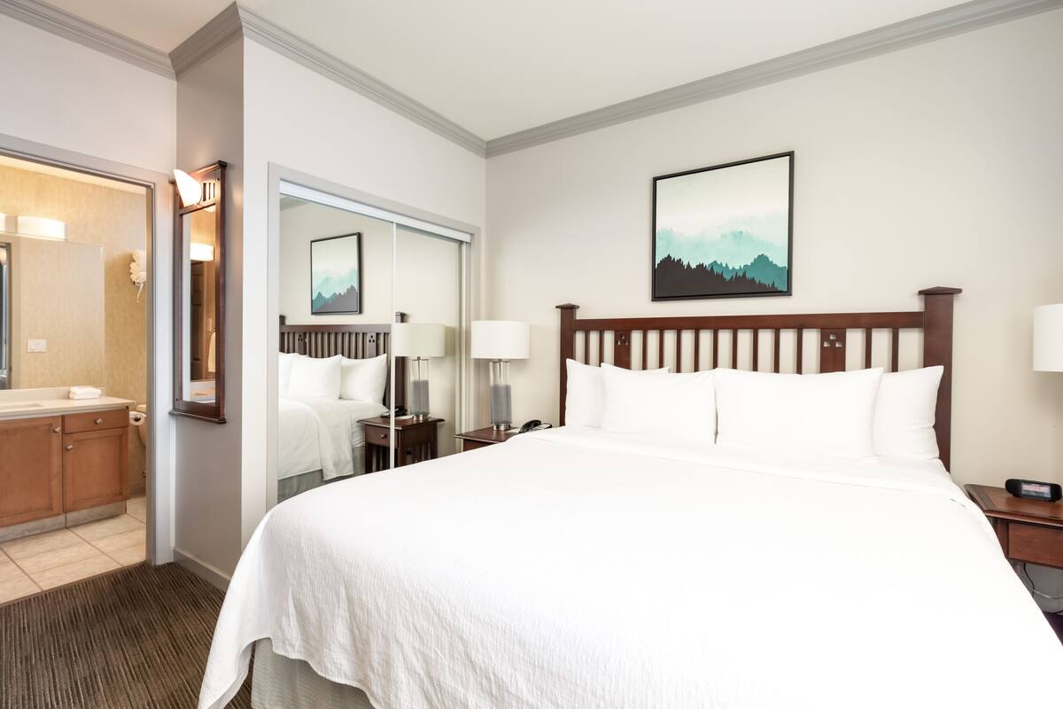 Habitación de hotel con una cama grande, un cabecero de madera, una mesita de noche con lámpara, una mesita auxiliar, un espejo y un cuadro en la pared.